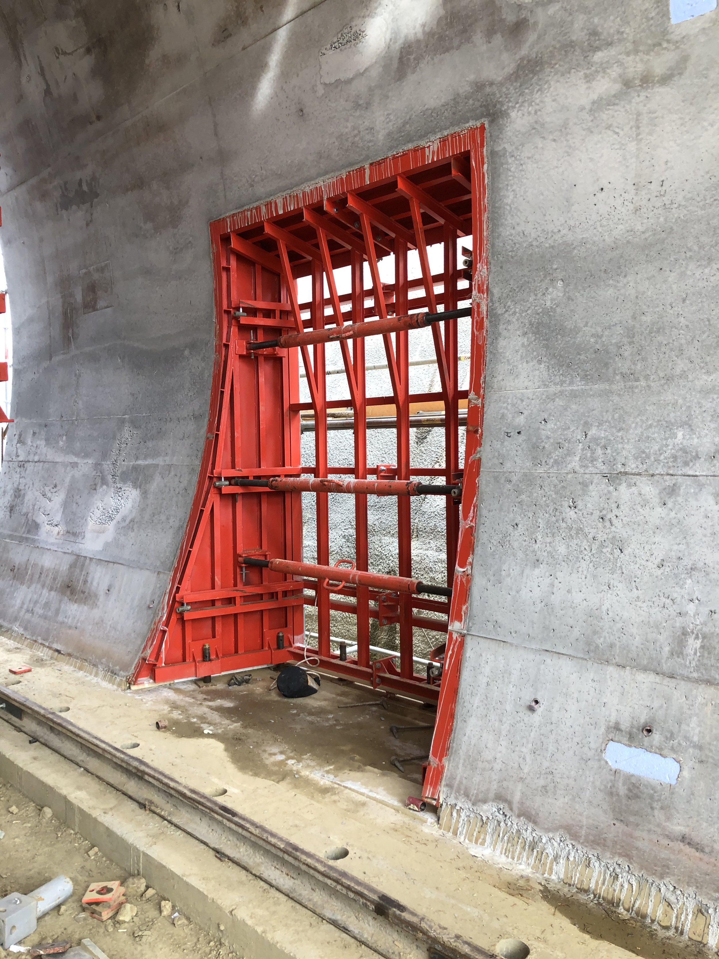 Construction site with concrete wall and red formwork for a window opening, supported by metal scaffolding, surrounding railroad tracks on the ground.