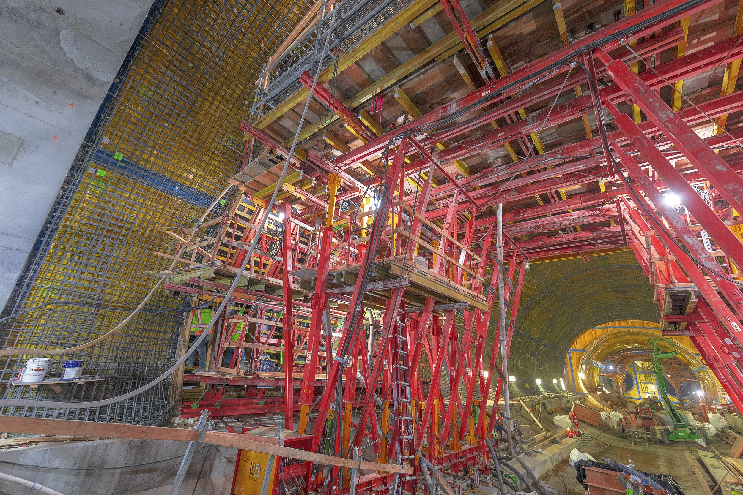 Interior view of a tunnel under construction with red scaffolding and yellow reinforcement mesh.