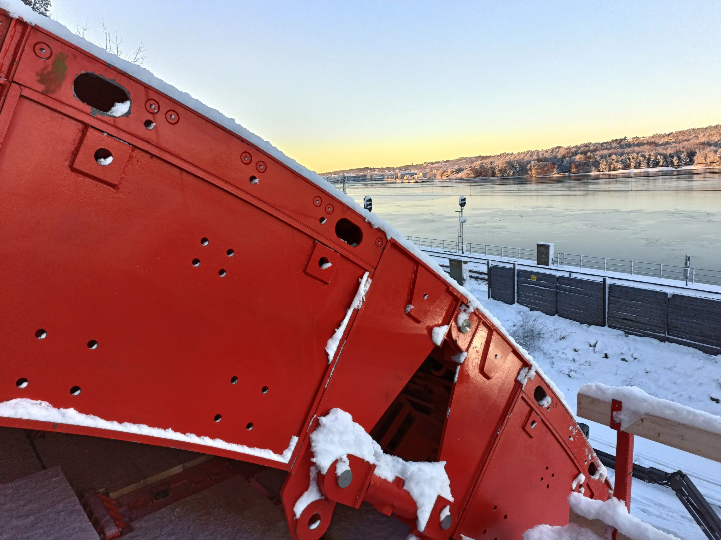 Close-up of a red snowplow blade with snow on it, set against a frozen river and a wintery landscape at sunrise.