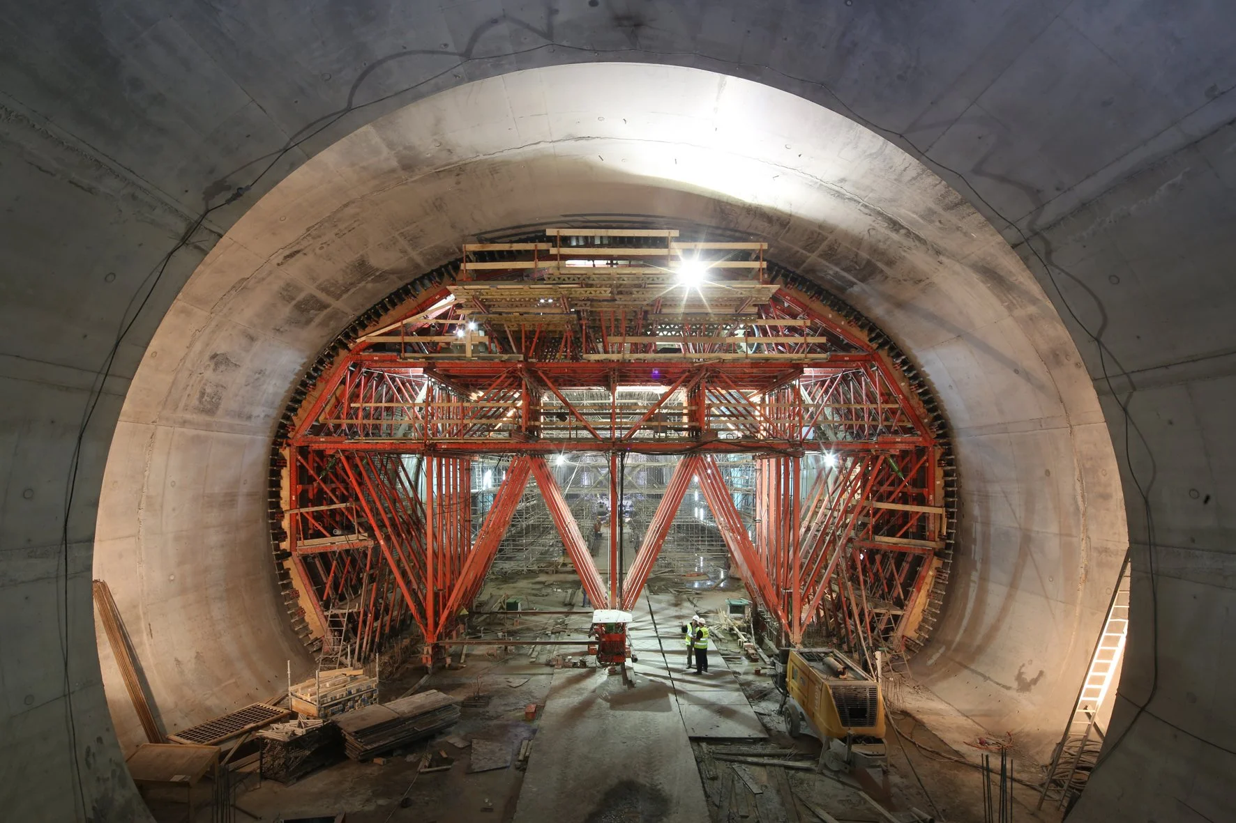 Construction workers inside a large underground tunnel with red scaffolding and concrete walls.