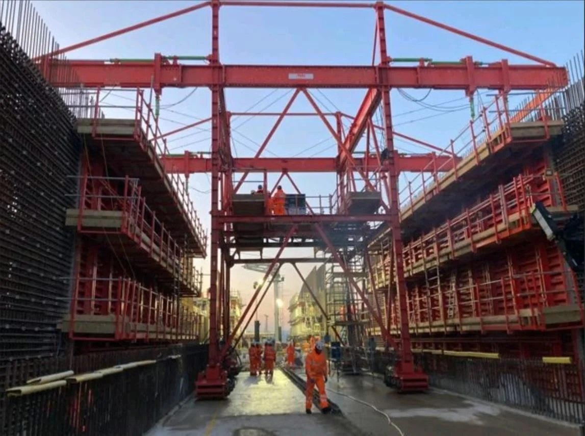 Construction workers wearing orange safety gear working on a large red steel structure at a construction site during daytime.