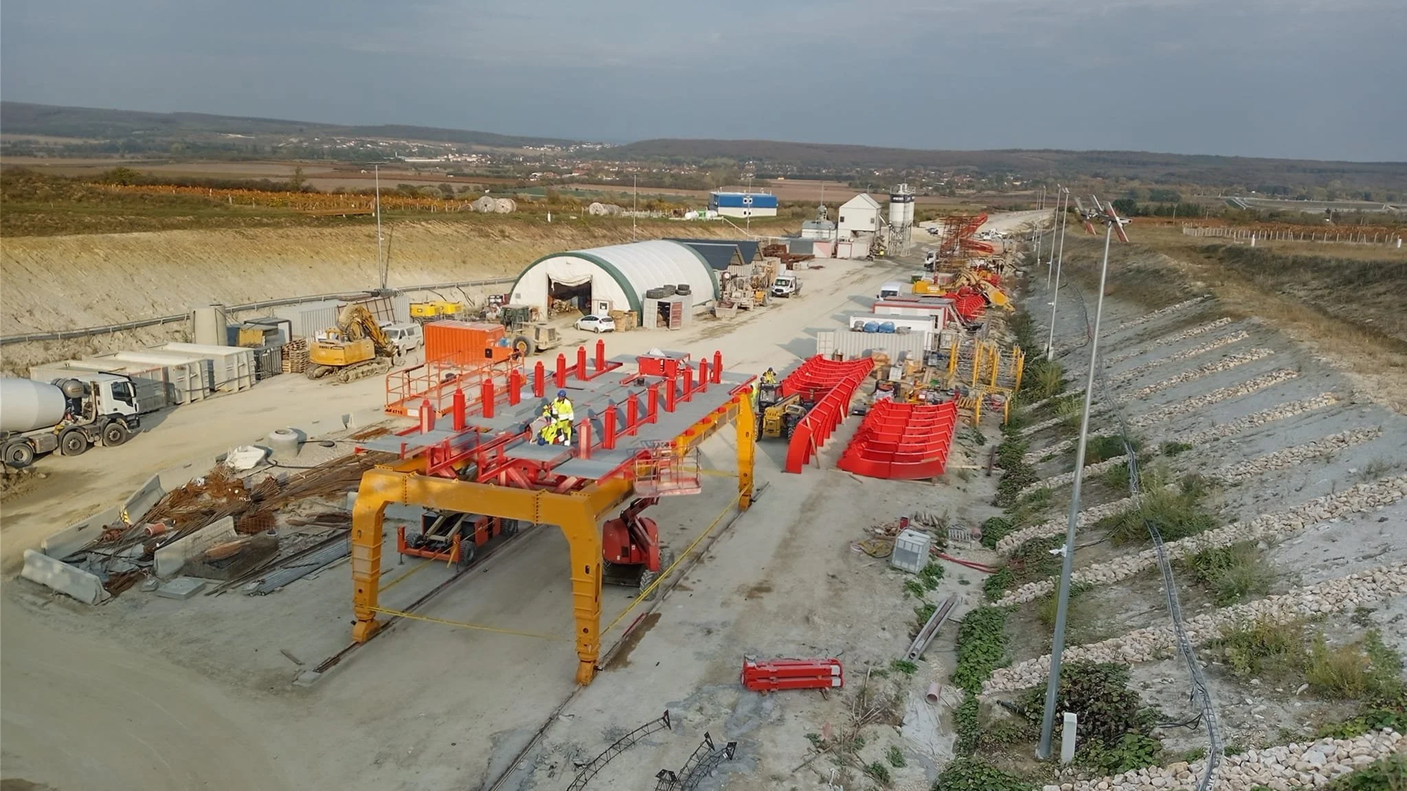Construction site with heavy machinery, workers, and red equipment being assembled or installed, adjacent to a roadway with hills in the background.