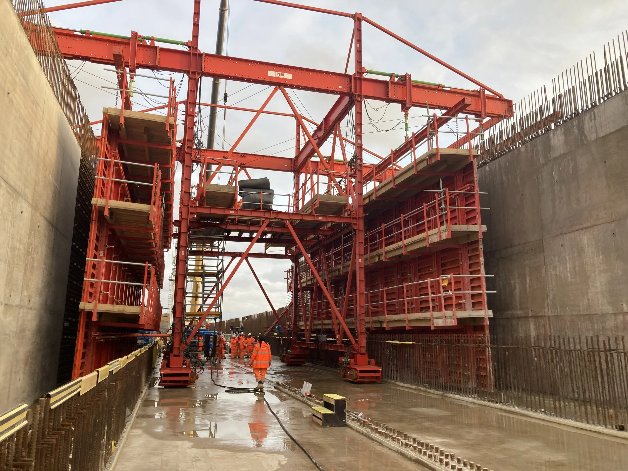Construction workers in orange safety uniforms working on a large construction site with a red scaffolding structure, concrete walls, and wet ground.