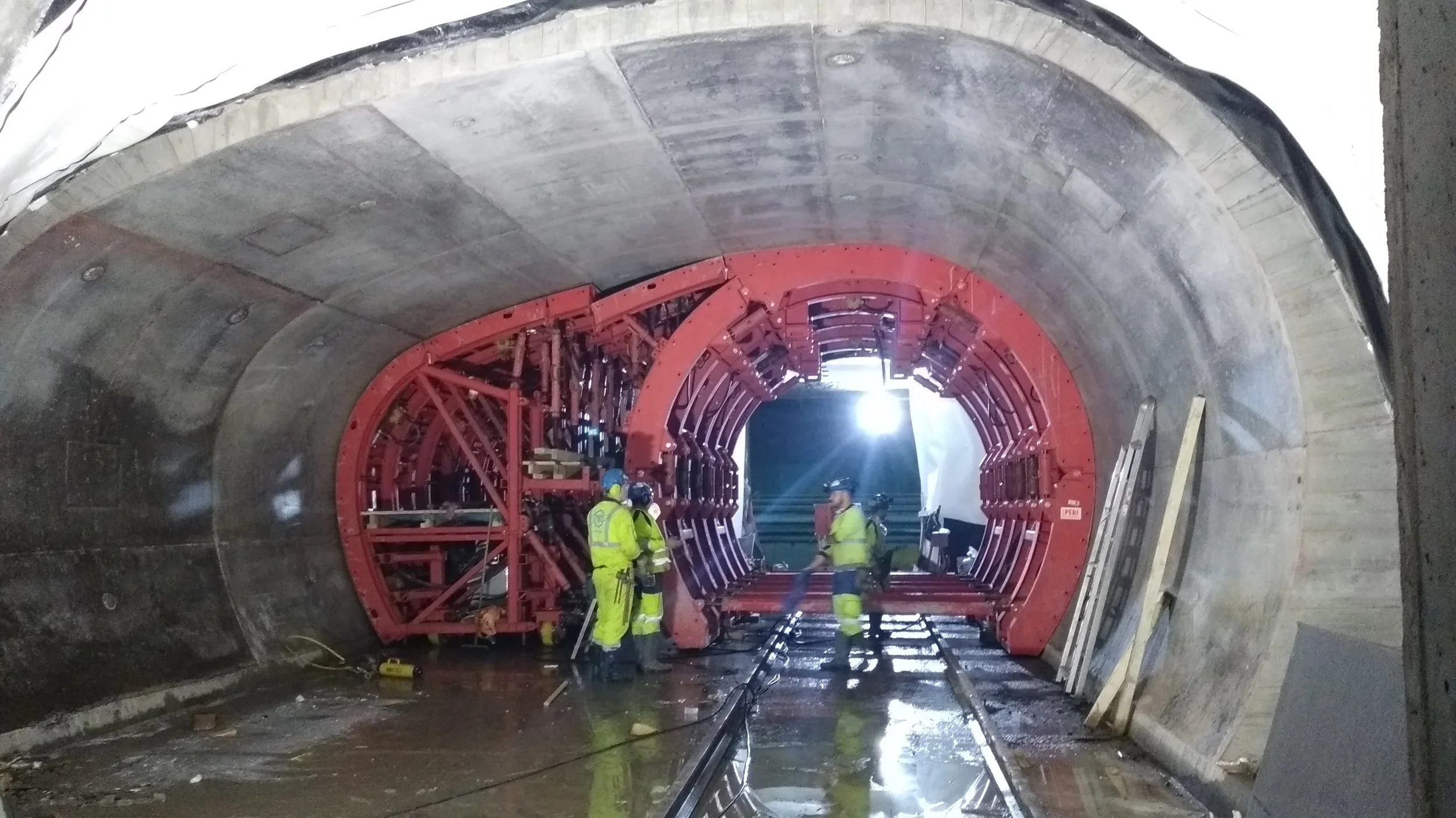 Construction workers in high-visibility clothing and helmets working inside a large tunnel with concrete walls and a red tunnel boring machine.