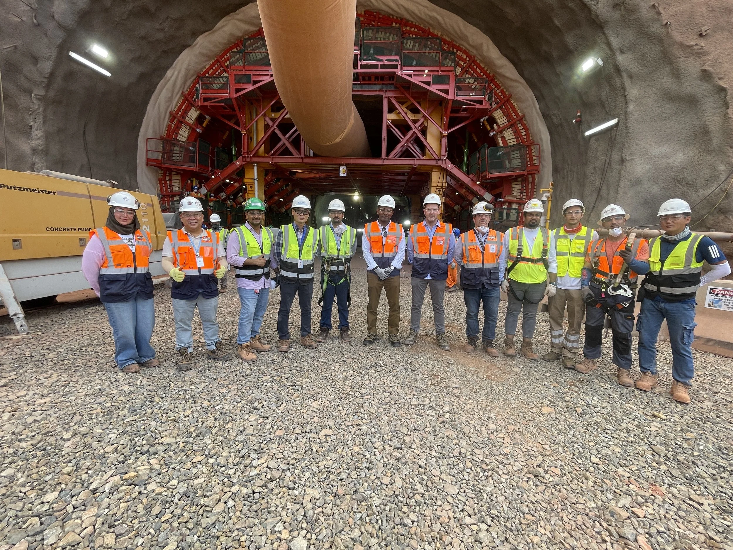 Group of construction workers wearing safety vests and helmets standing inside a tunnel with construction equipment and scaffolding.