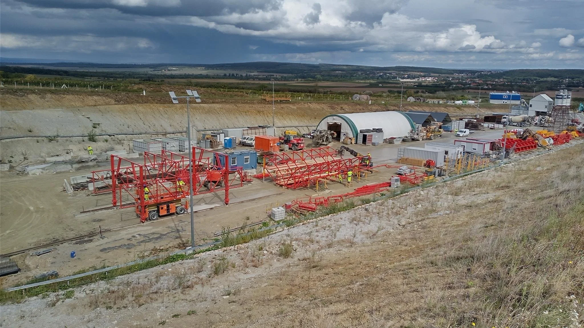 Construction site with red steel framework, construction workers, and various equipment, set in a rural area with cloudy sky.