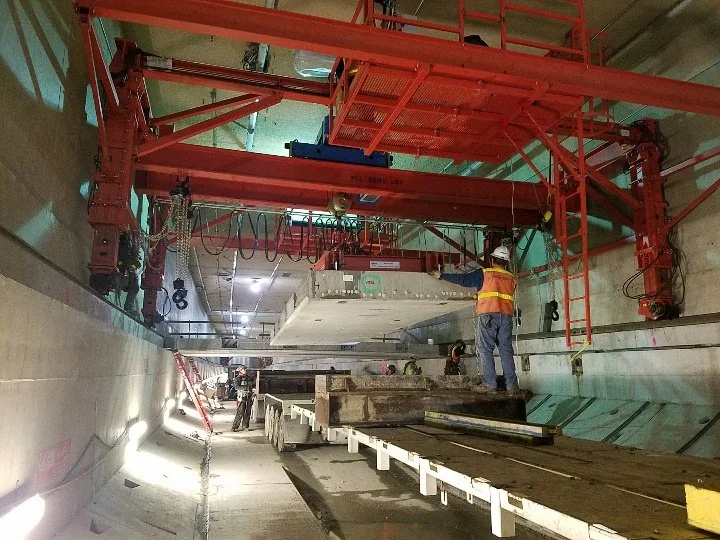 Construction workers guide a large concrete section being lowered into an underground tunnel as part of tunnel or metro construction project, with overhead machinery and scaffolding visible.