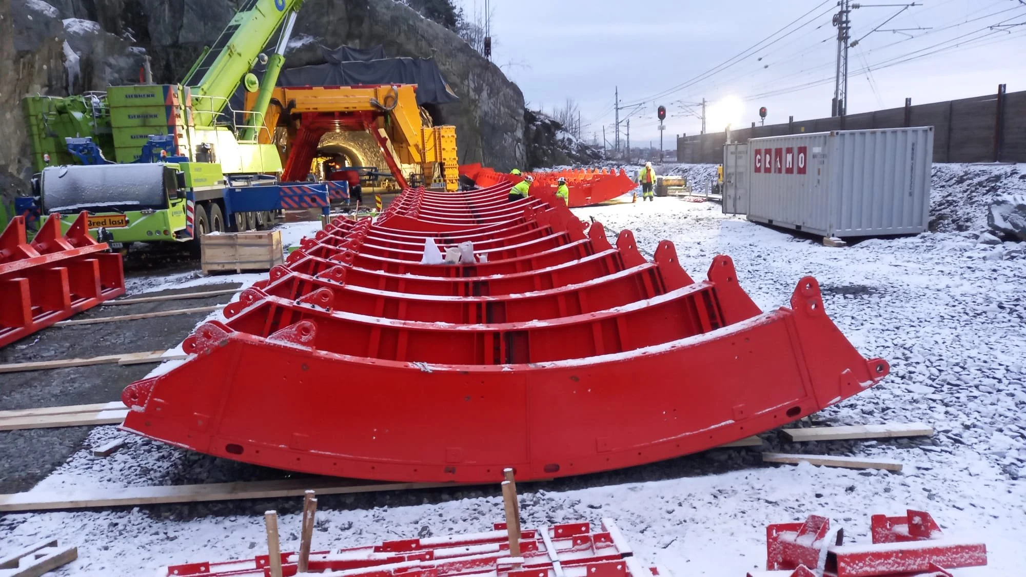 Construction workers installing large red tunnel sections on a snowy railway track with a crane, truck, and shipping containers in the background.