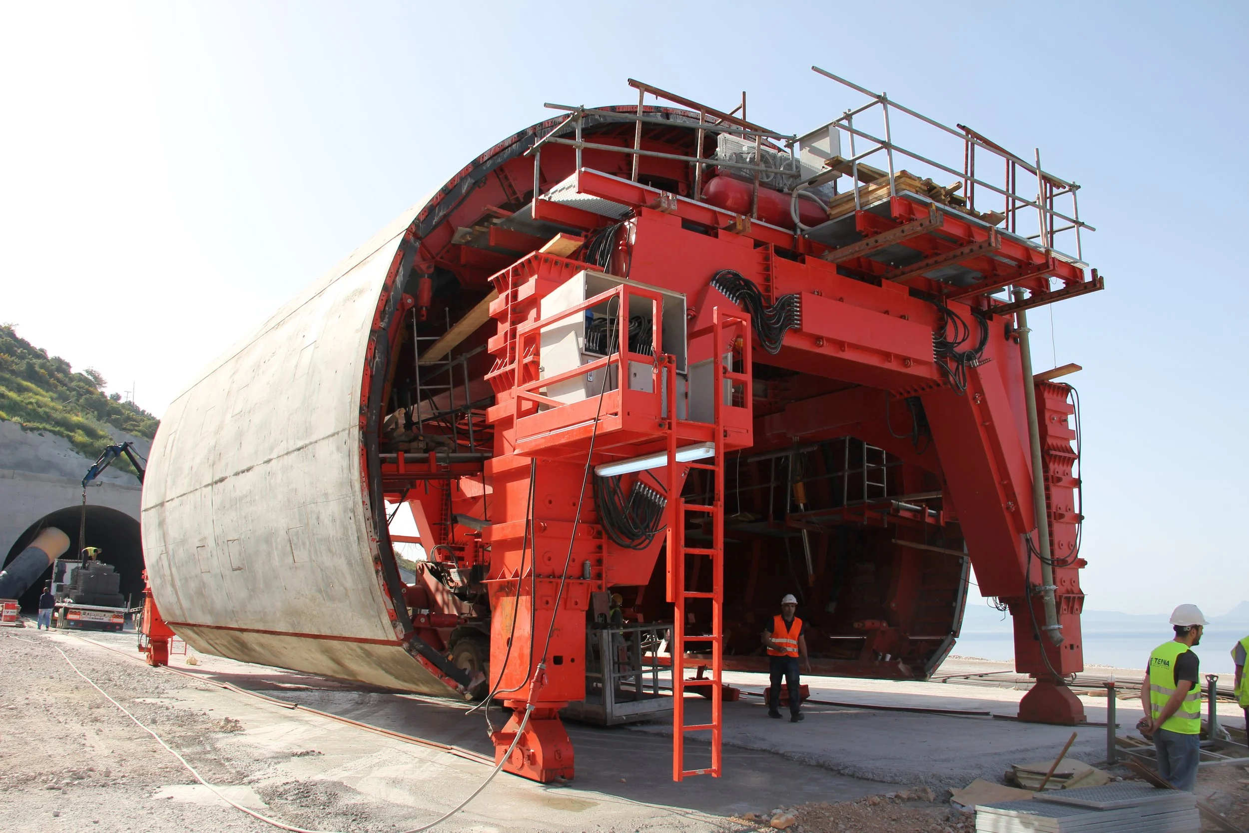 Large construction or engineering equipment with a circular shape, red frame, and various attached components, situated outdoors with workers in safety vests and helmets nearby.