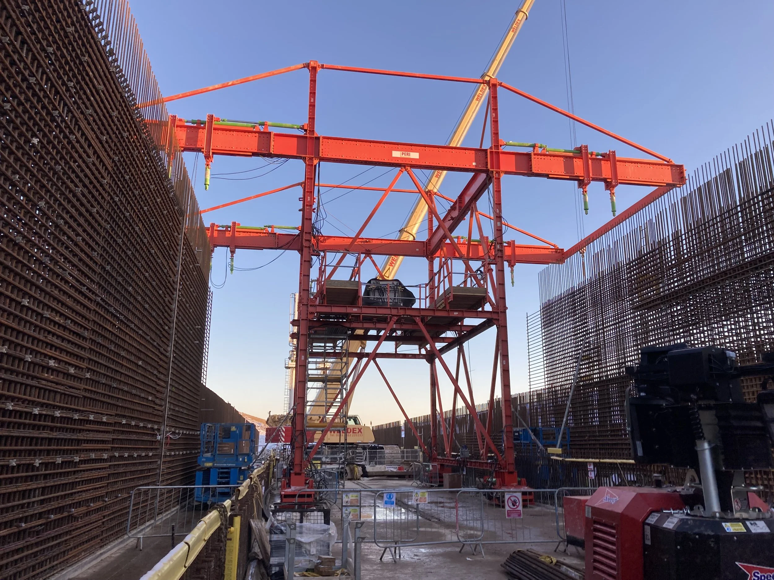 Construction site with a large red crane and tall brown metal barriers on either side, under a clear blue sky.