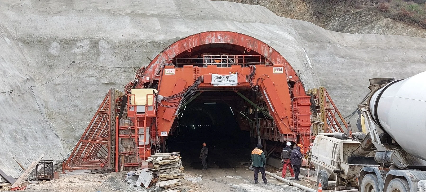 Workers standing in front of a tunnel construction site with a large red tunnel boring machine, surrounded by construction equipment and dirt.