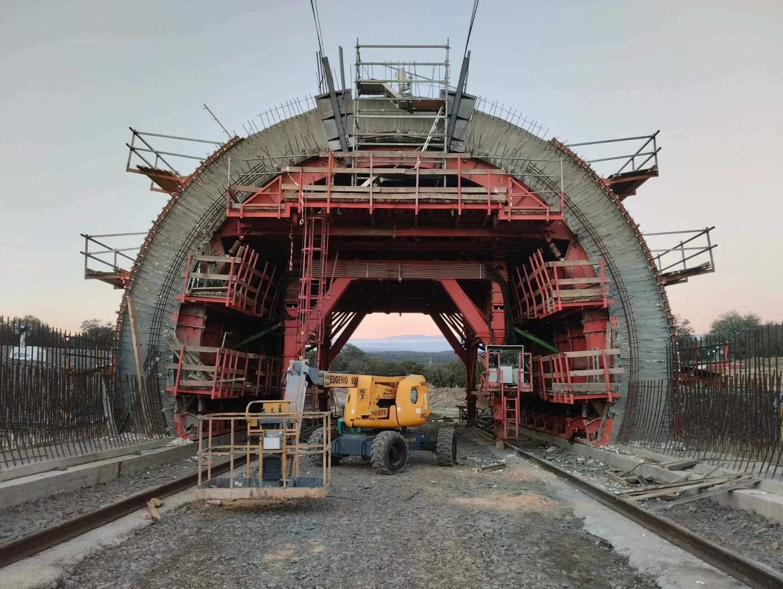 Construction site of a large tunnel with steel framework and safety railings, with a yellow lift in the foreground and railroad tracks on either side.