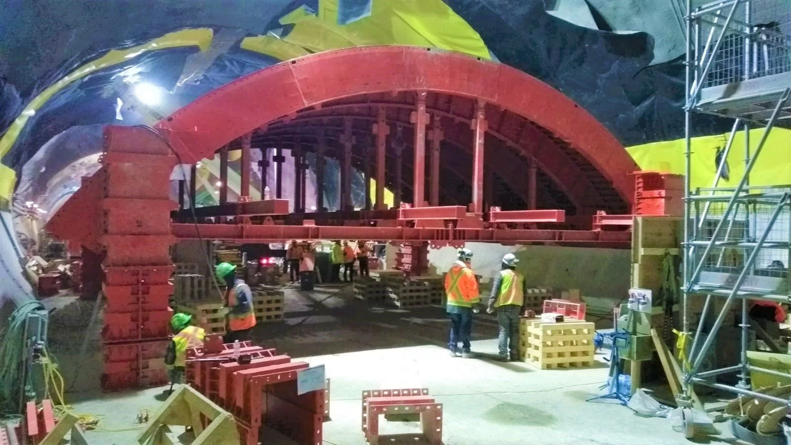 Construction workers in helmets and safety vests at a building site inside a tunnel, assembling large red steel arches for tunnel support.