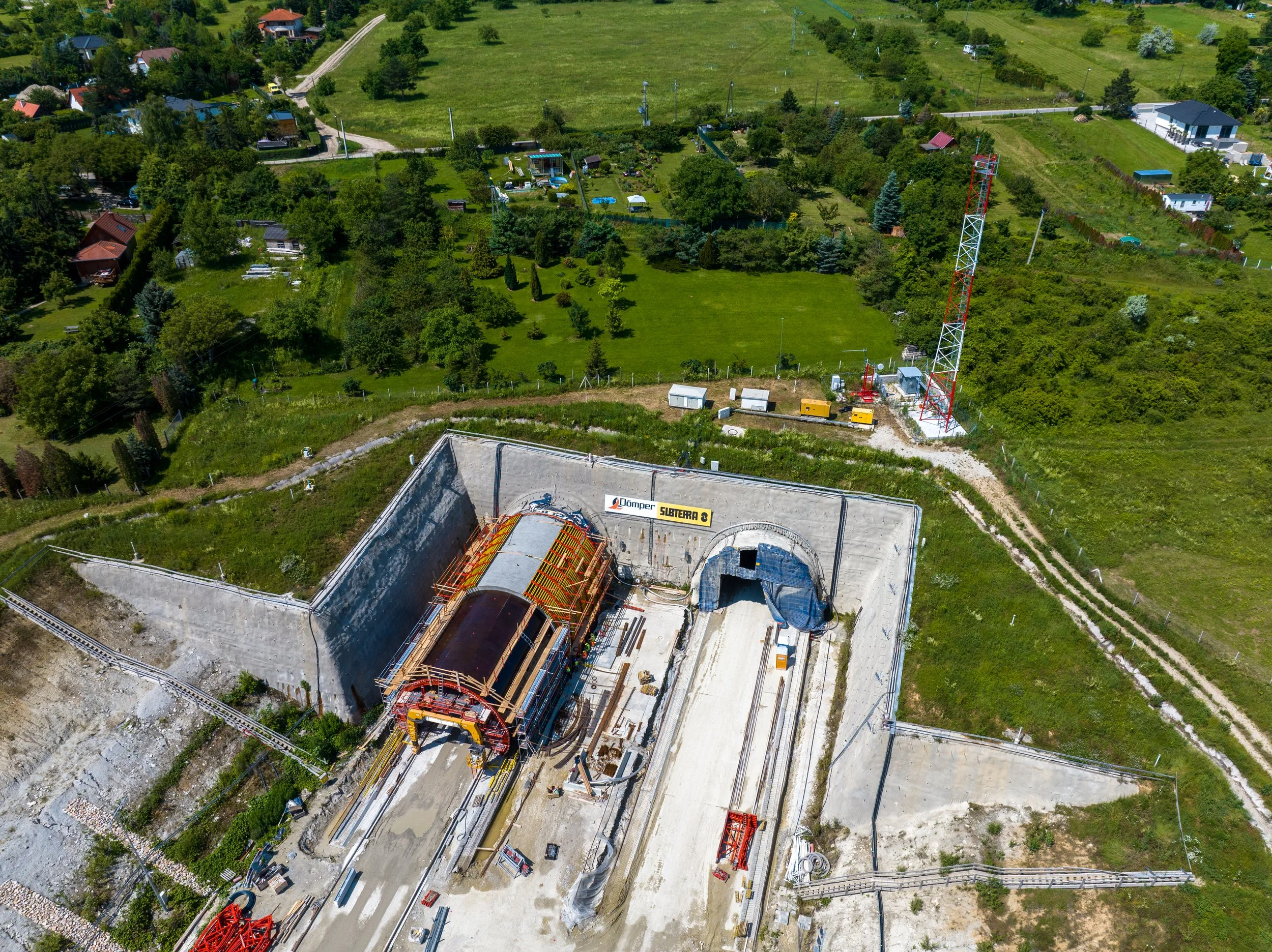 Aerial view of a construction site for a train tunnel with construction equipment, a large tunnel entrance, and a railway track, surrounded by greenery and residential houses.