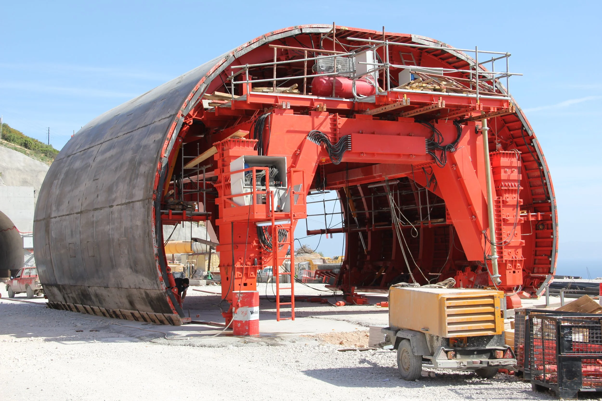 A large, red industrial machine under construction, resembling a tunnel boring machine, with scaffolding and various equipment around it at a construction site.
