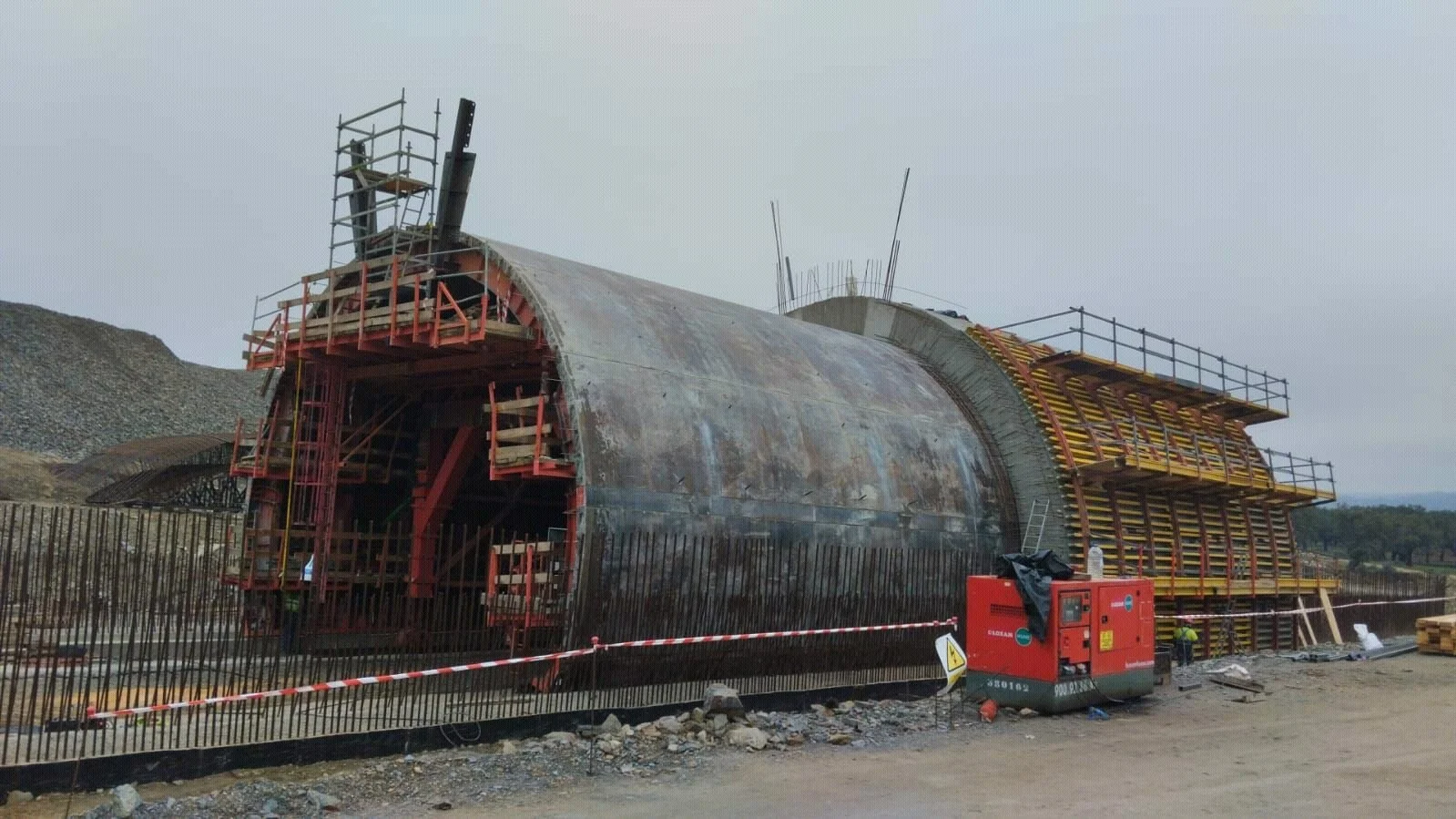 Construction site with a large cylindrical metal structure and scaffolding. Workers are visible, and equipment is positioned around the site.