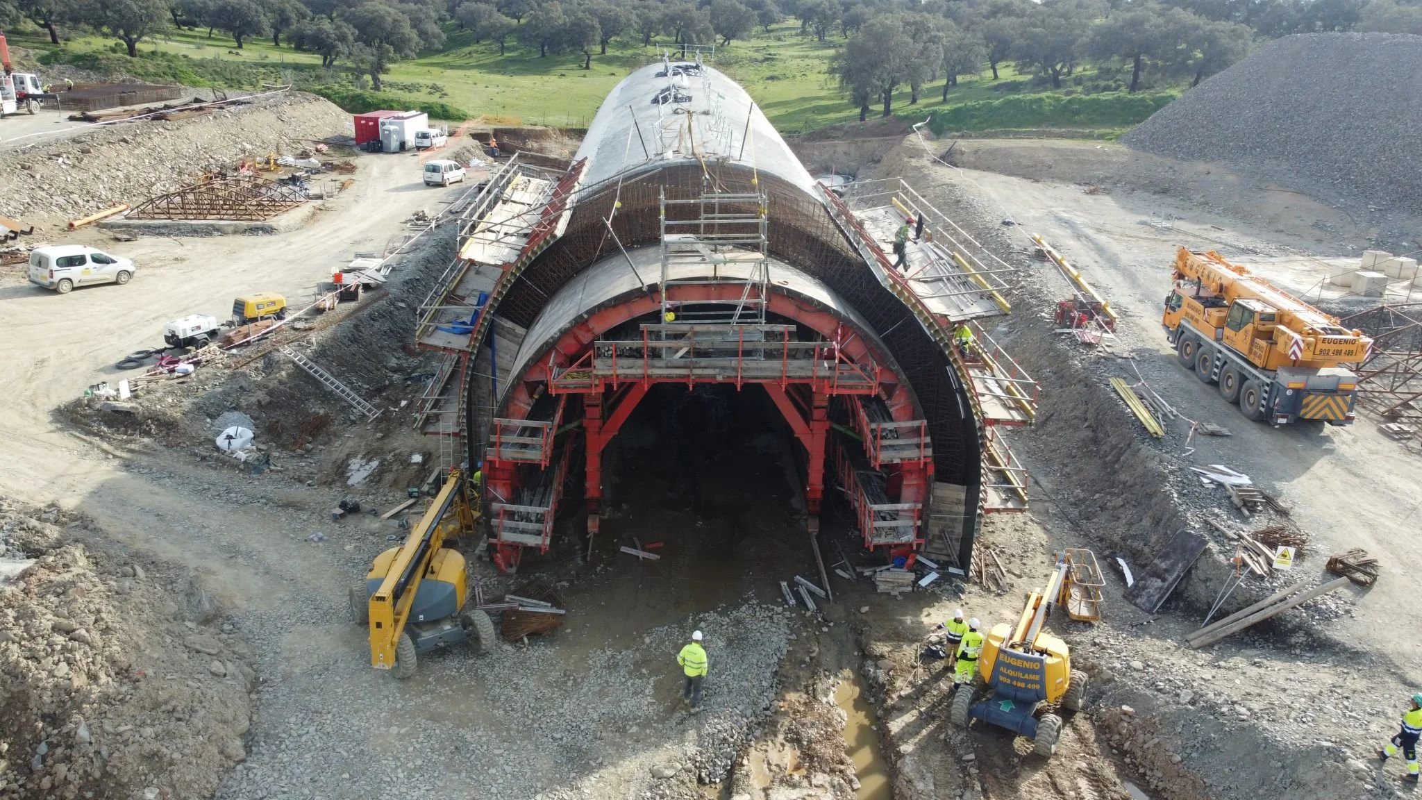 Construction site with a tunnel being built, workers, and construction equipment, including cranes and excavators, at a rural location.