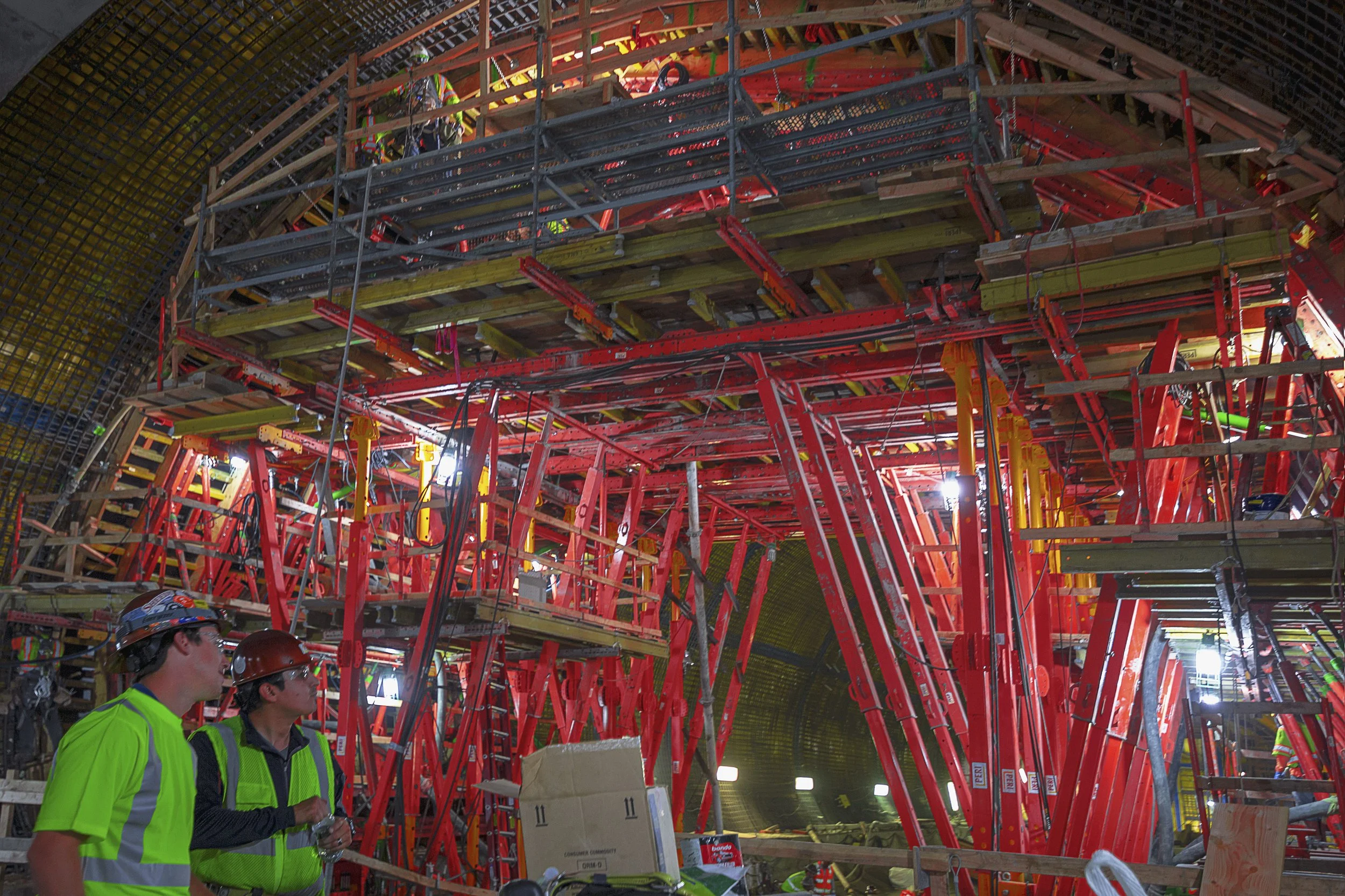 Construction workers in safety gear standing inside a large tunnel with complex red and yellow scaffolding and support structures.