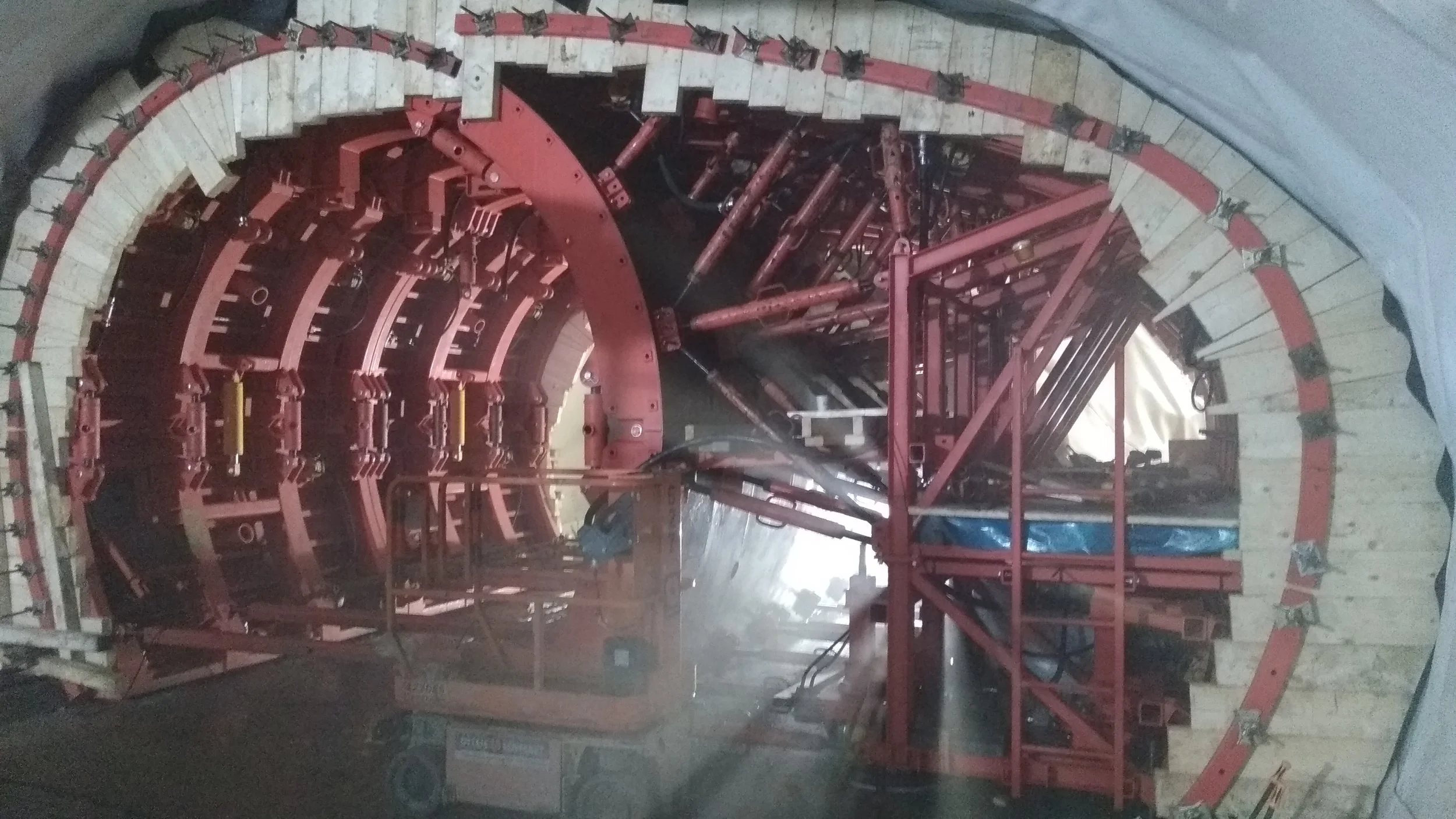 A tunnel boring machine with red and beige components, including a large, circular cutting head and mechanical arms, inside a tunnel under construction.