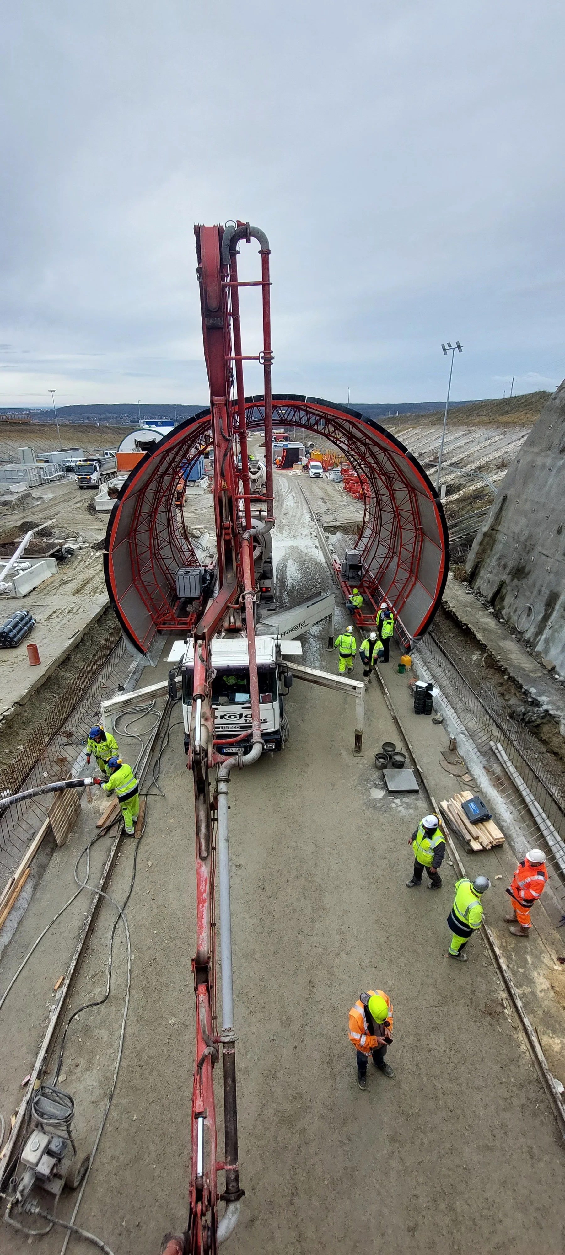 Construction workers on a road construction site, pouring concrete with large machinery and a curved tunnel structure in the background.