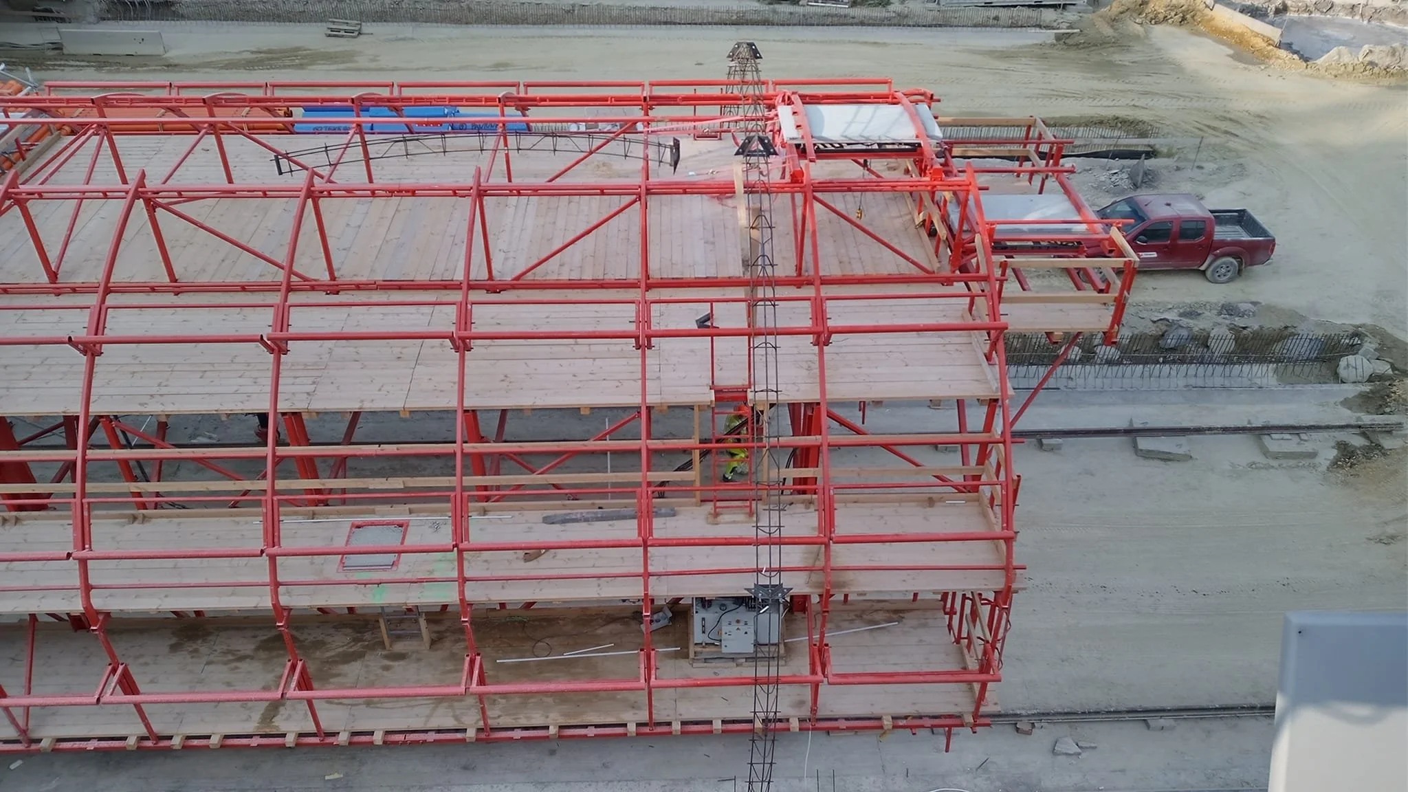 Aerial view of a multi-level construction site with red steel framing and wooden flooring, a worker in safety gear, and a parked red pickup truck on a dirt road.