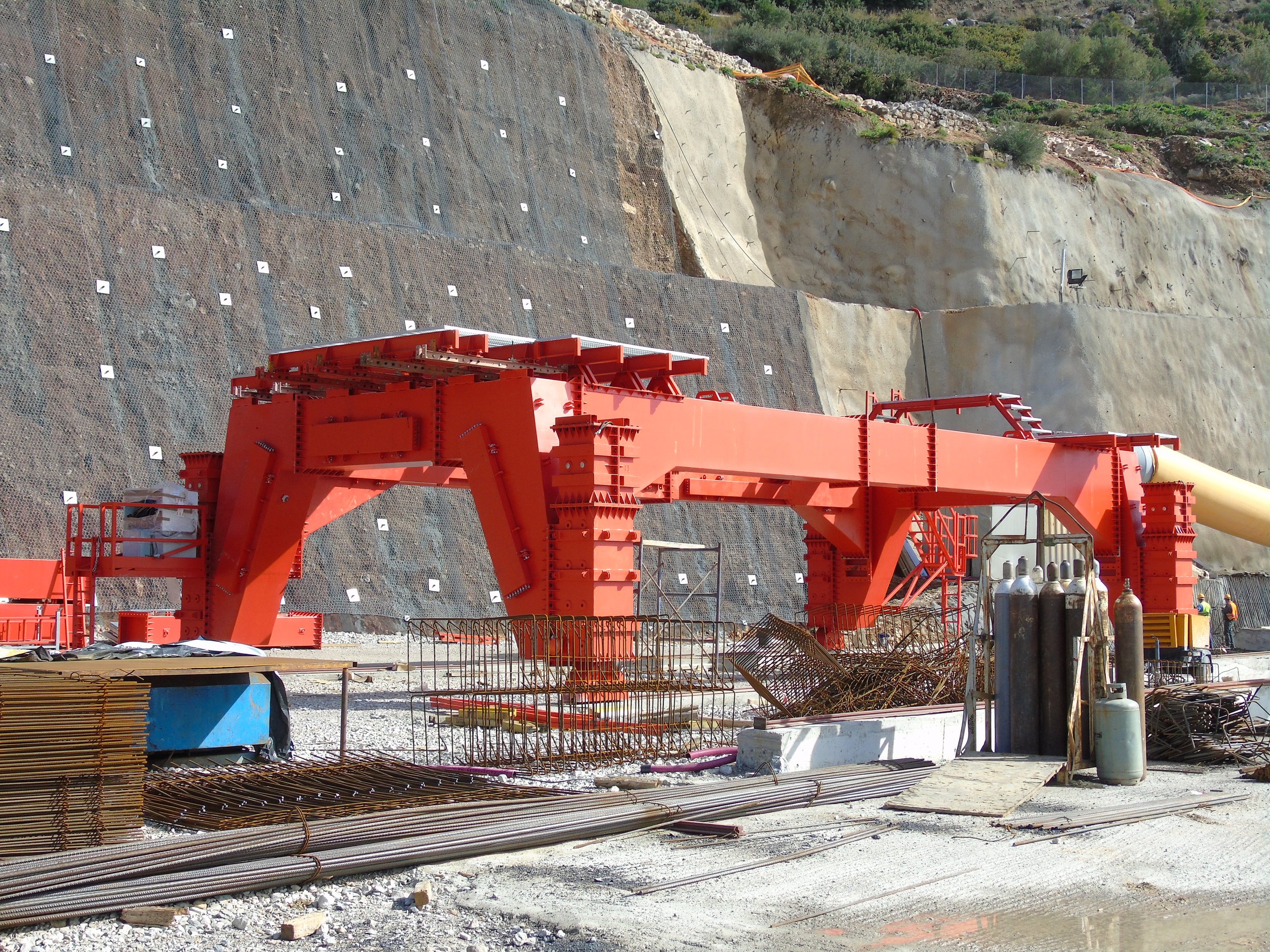 Large red construction machine at a construction site with gravel, rebar, and gas tanks in the foreground, and a hillside with netting and rocks in the background.