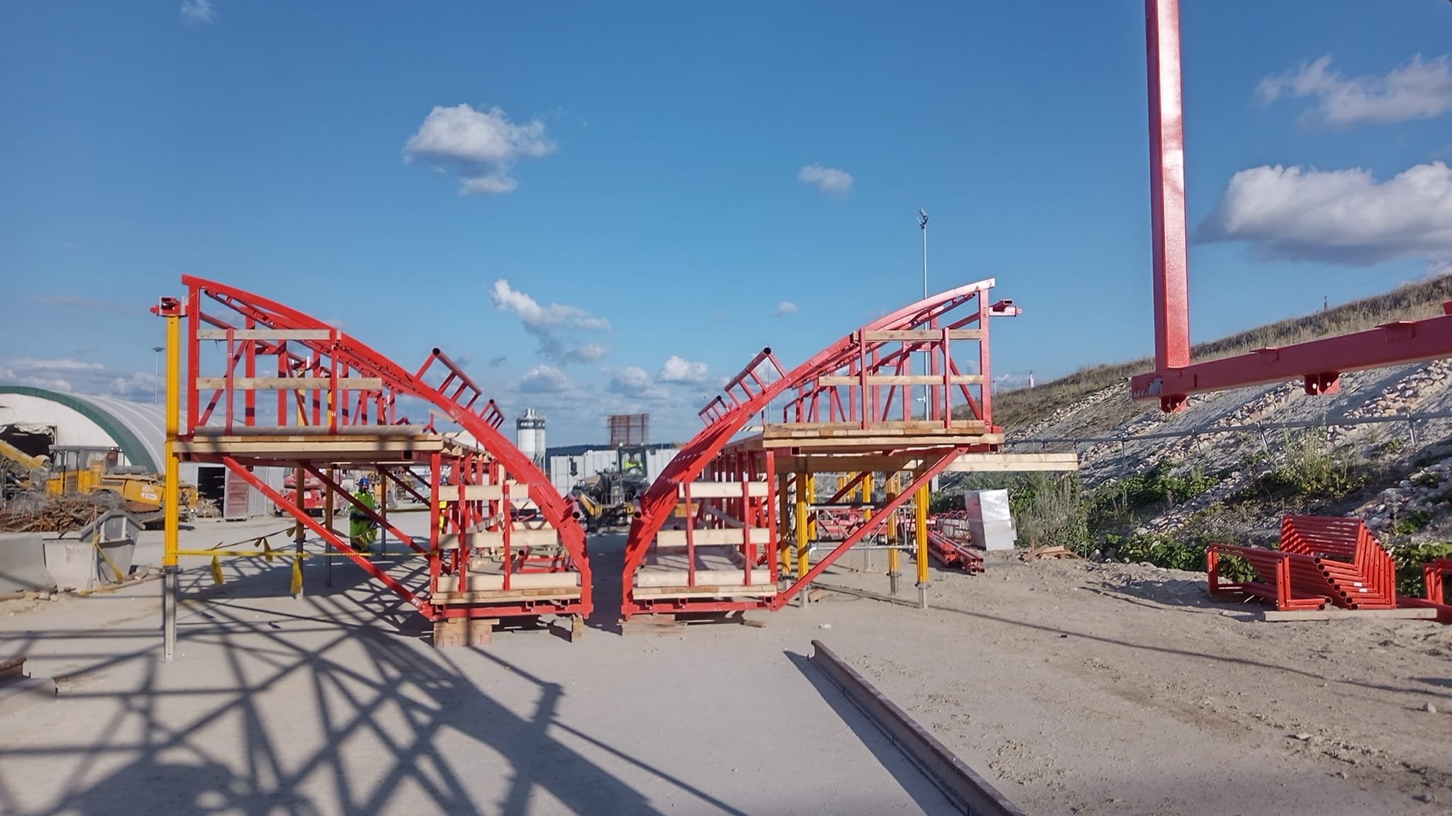 Construction site with red steel framework being assembled on a dirt ground, with blue sky and clouds in the background.