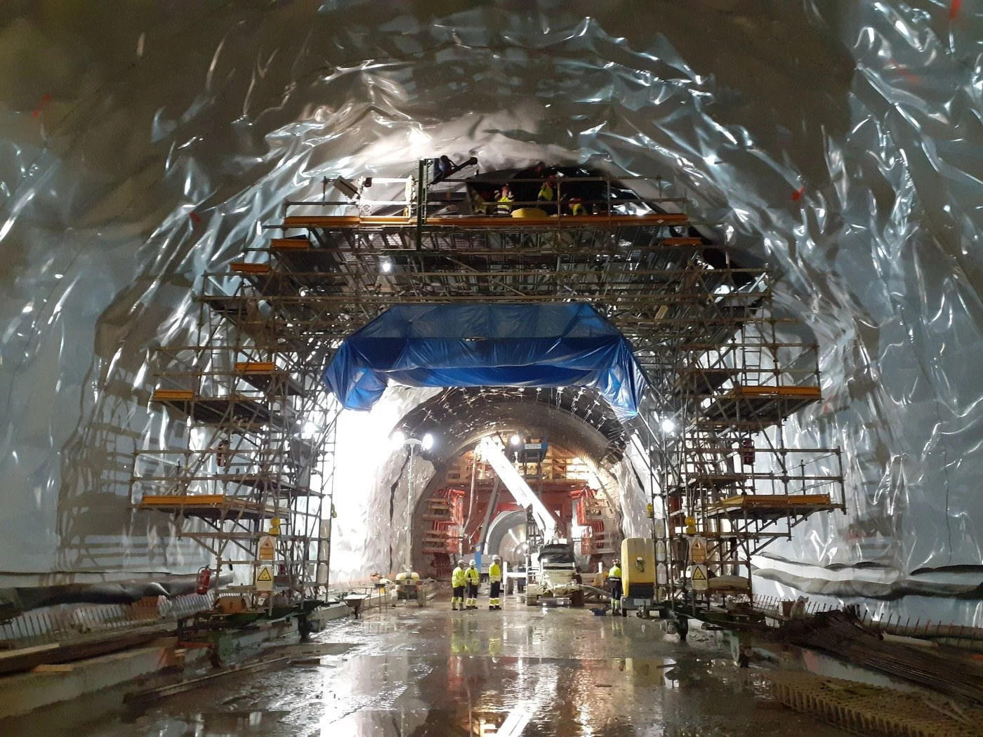 Construction workers working on a tunnel under construction, with scaffolding, equipment, and reflective wall lining.