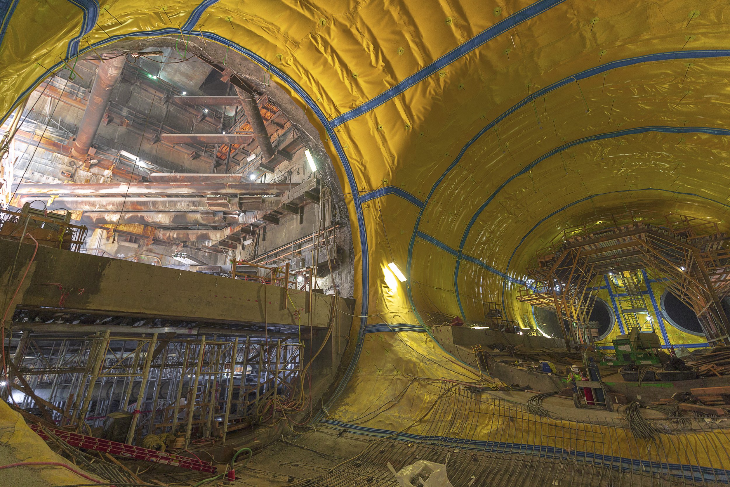 Interior of a large underground construction tunnel with yellow and blue inflatable or protective lining, construction scaffolding, and steel pipes.