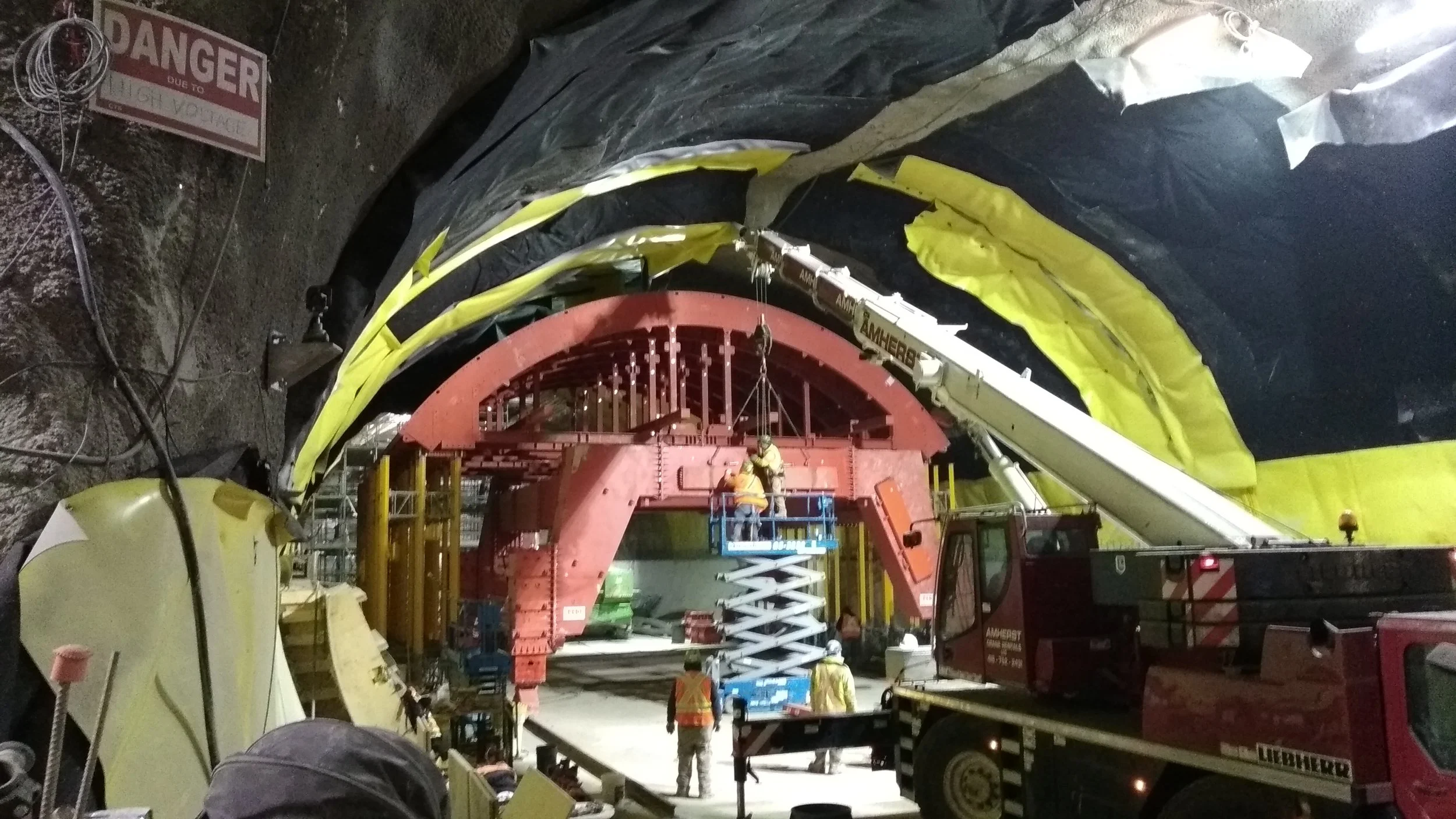Construction workers operating a crane and lift inside a large tunnel, with a red segment structure and safety equipment, and a 'Danger' warning sign on the wall.