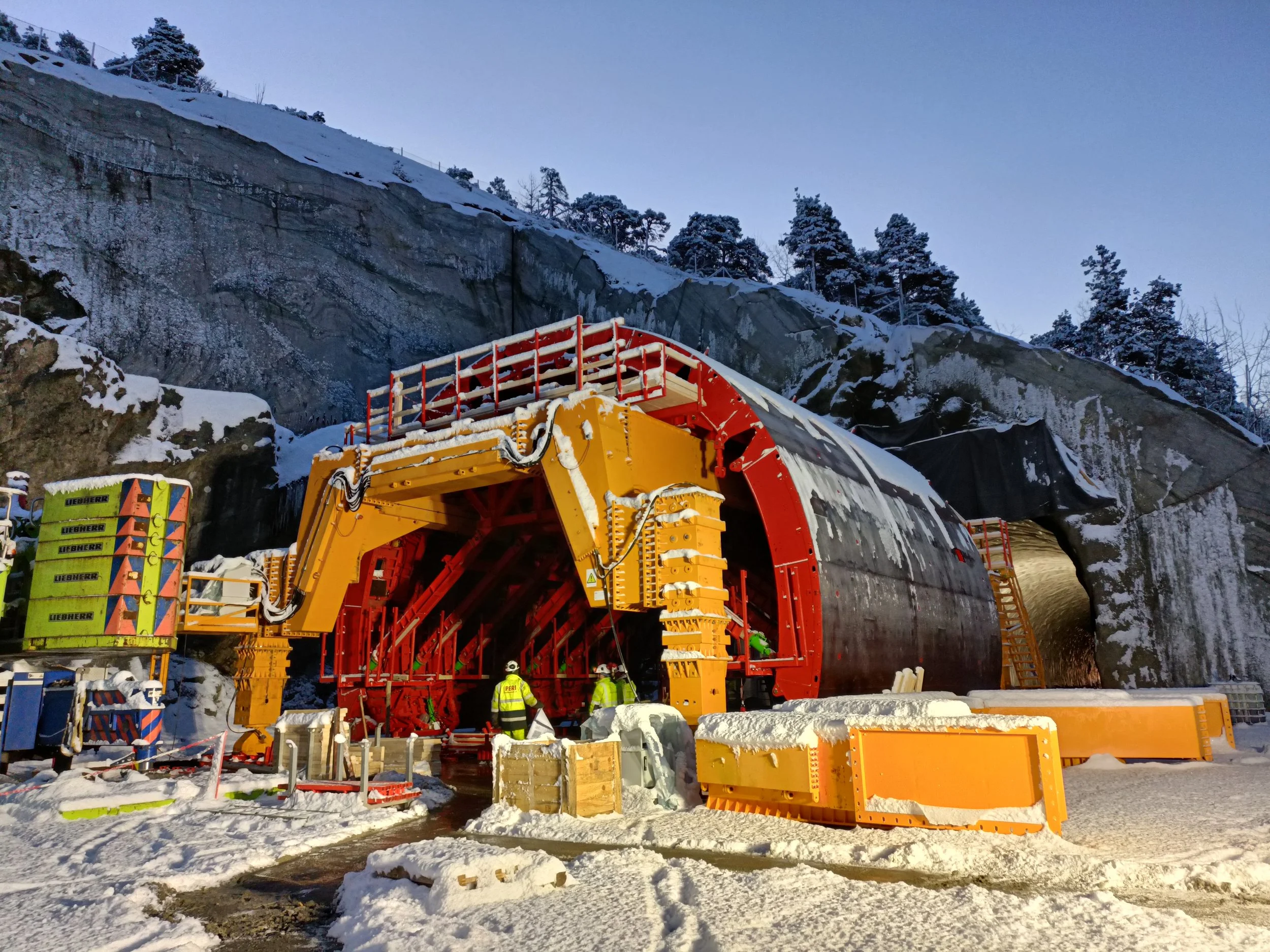Construction site of a tunnel with snowy landscape, workers in safety gear, and large orange and yellow tunnel boring machine under a rocky and snow-covered hillside.