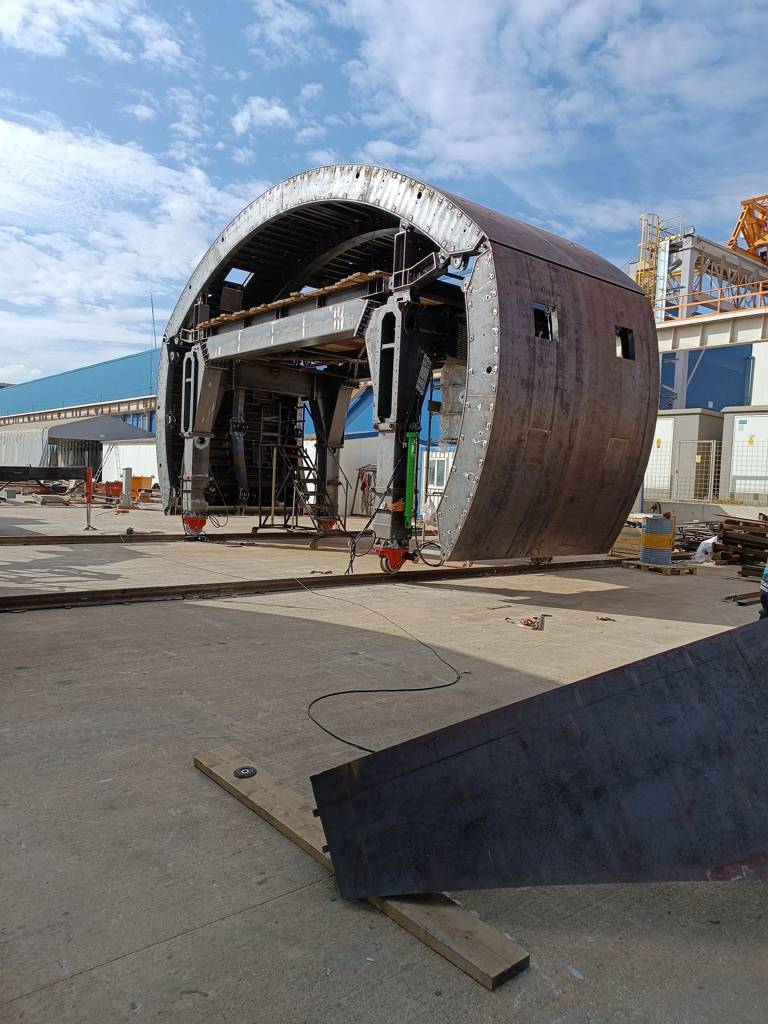 Large metal structure under construction, resembling a spacecraft or large industrial equipment, situated outdoors on a concrete surface with tools and materials nearby. The background includes industrial buildings and a partly cloudy sky.
