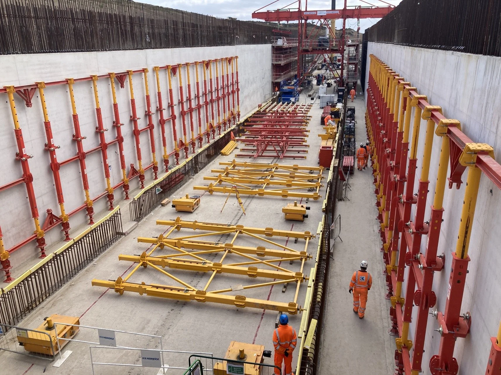 Construction workers in orange safety uniforms and helmets working inside a large, deep excavated construction site. The site has large yellow and red hydraulic shoring equipment installed along the walls for support, with some equipment laid out on 