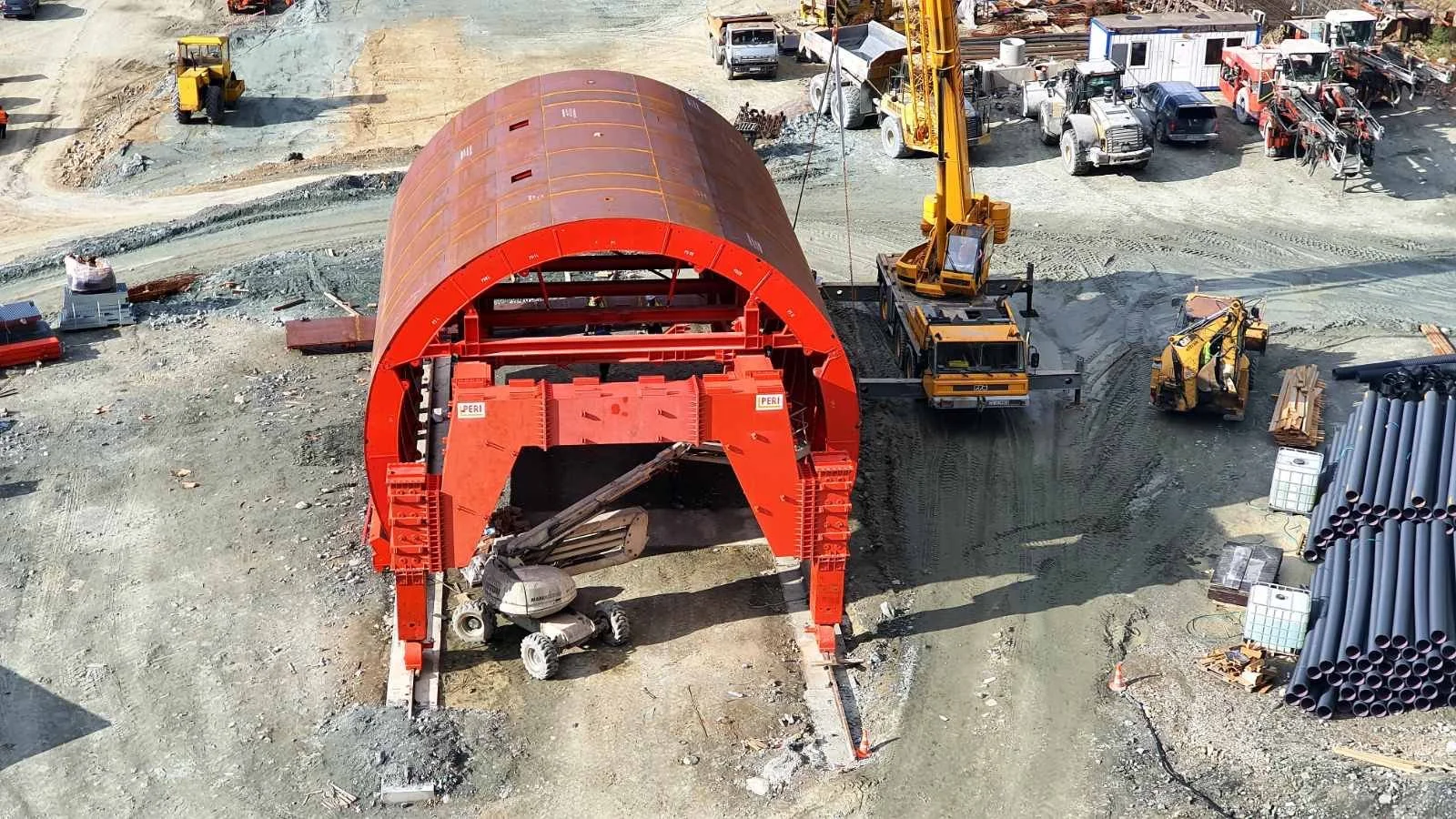 Construction site with a large red and brown tunnel structure, construction vehicles, pipes, and materials for building infrastructure.