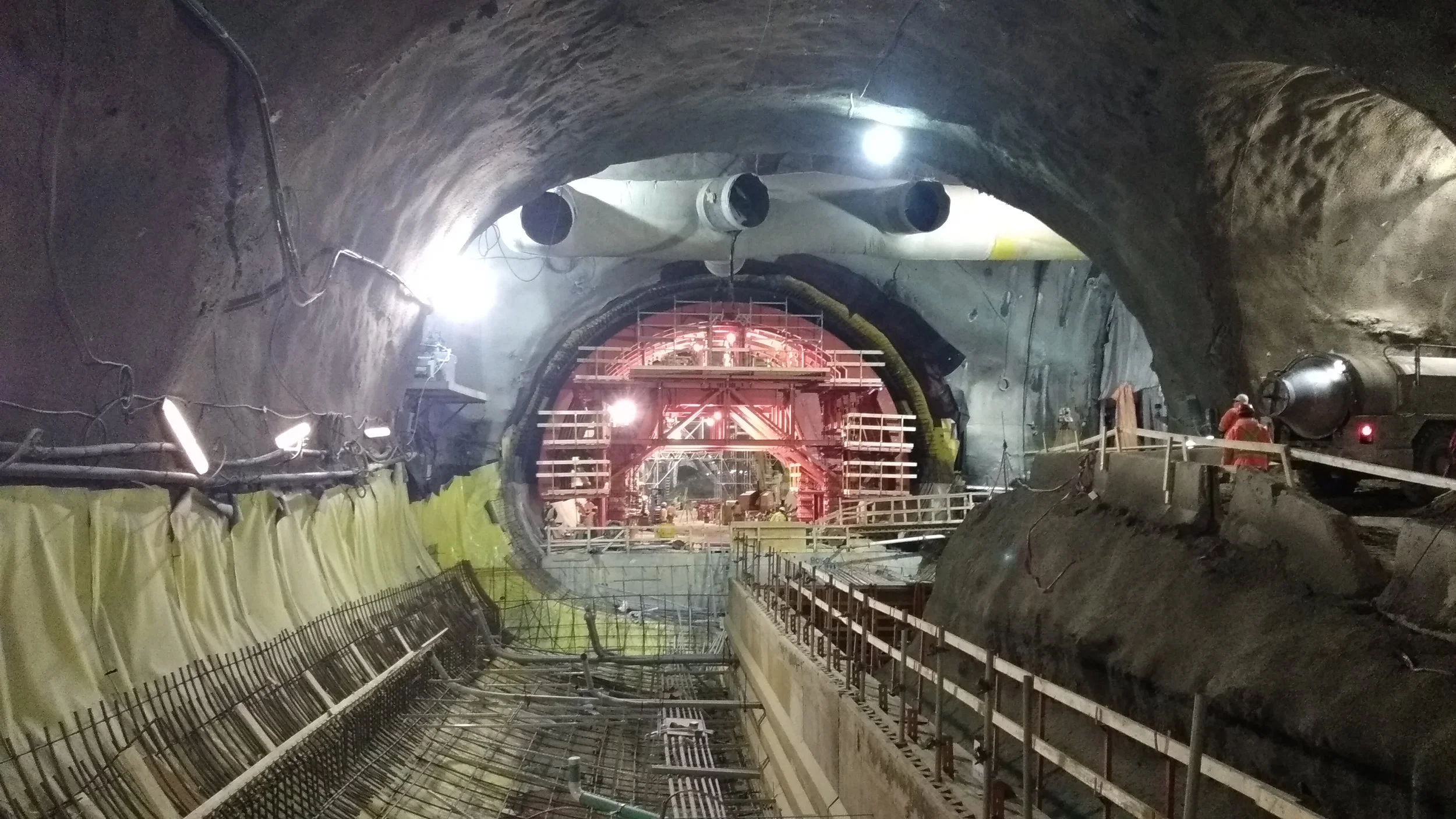 Underground construction site with workers, scaffolding, pipes, and lighting in a tunnel or large excavation area.