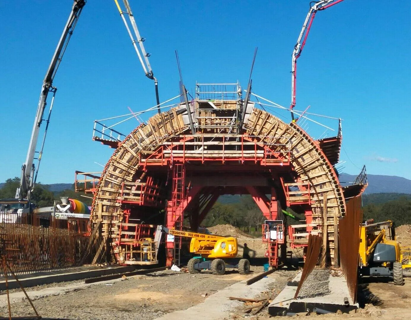 Construction site with a large tunnel-like structure made of wood and steel, surrounded by construction equipment, with a clear blue sky and distant mountains in the background.