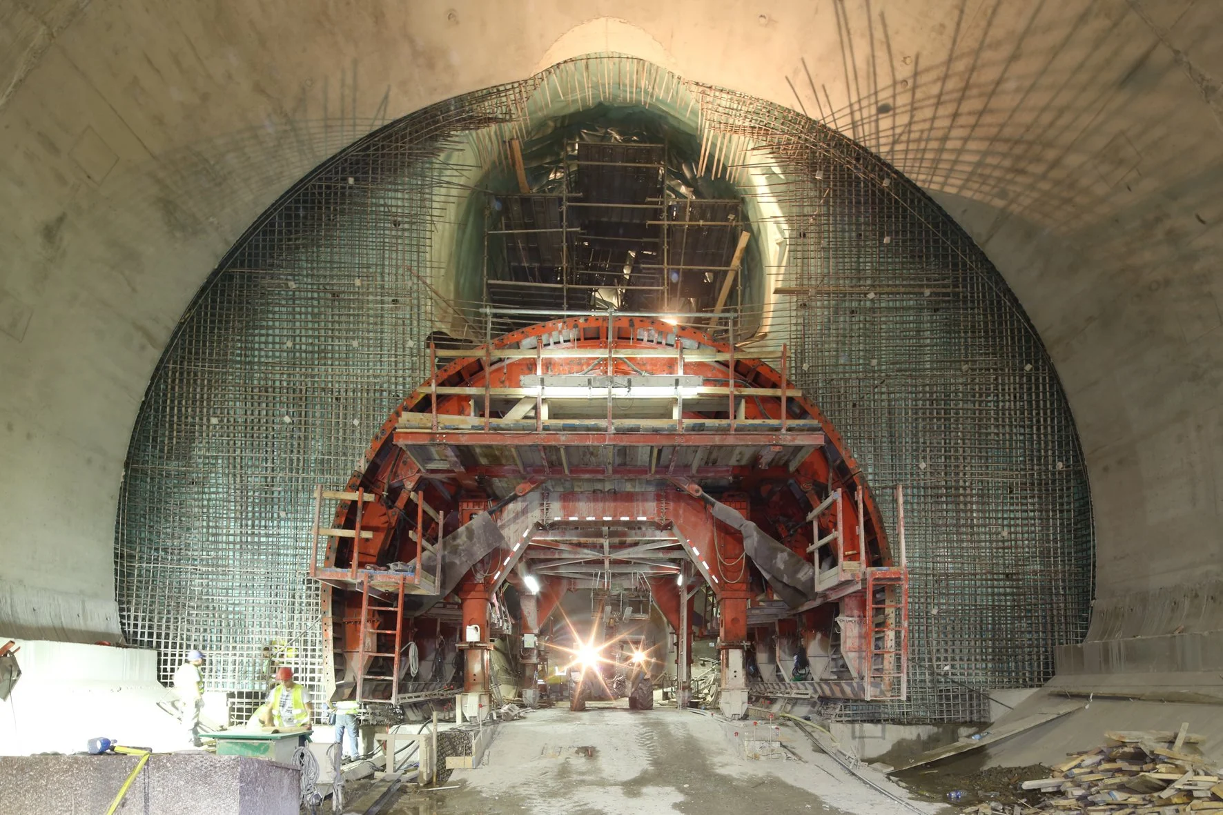 Construction workers working inside a tunnel with scaffolding and reinforced steel mesh. The tunnel is partially built with exposed concrete walls.