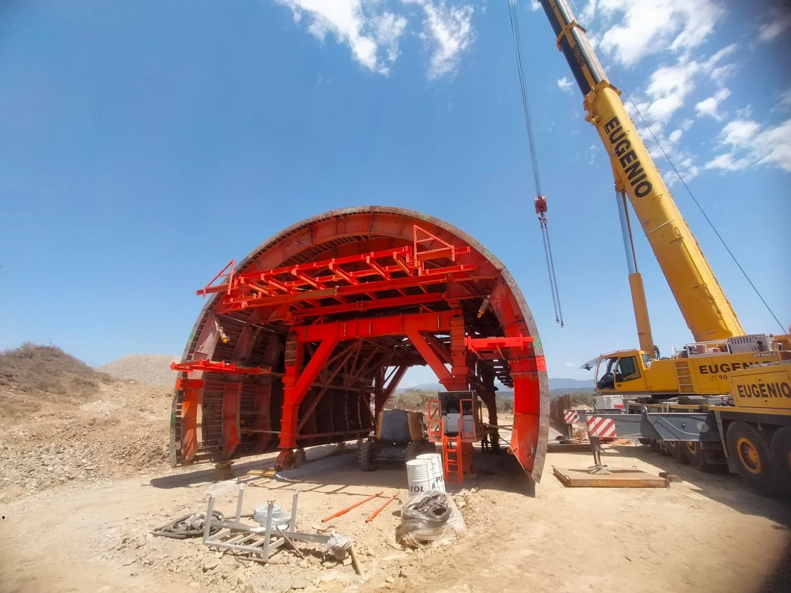 Construction site with a large tunnel segment and a crane. The segment is supported by scaffolding and structures inside, with a bright blue sky overhead.