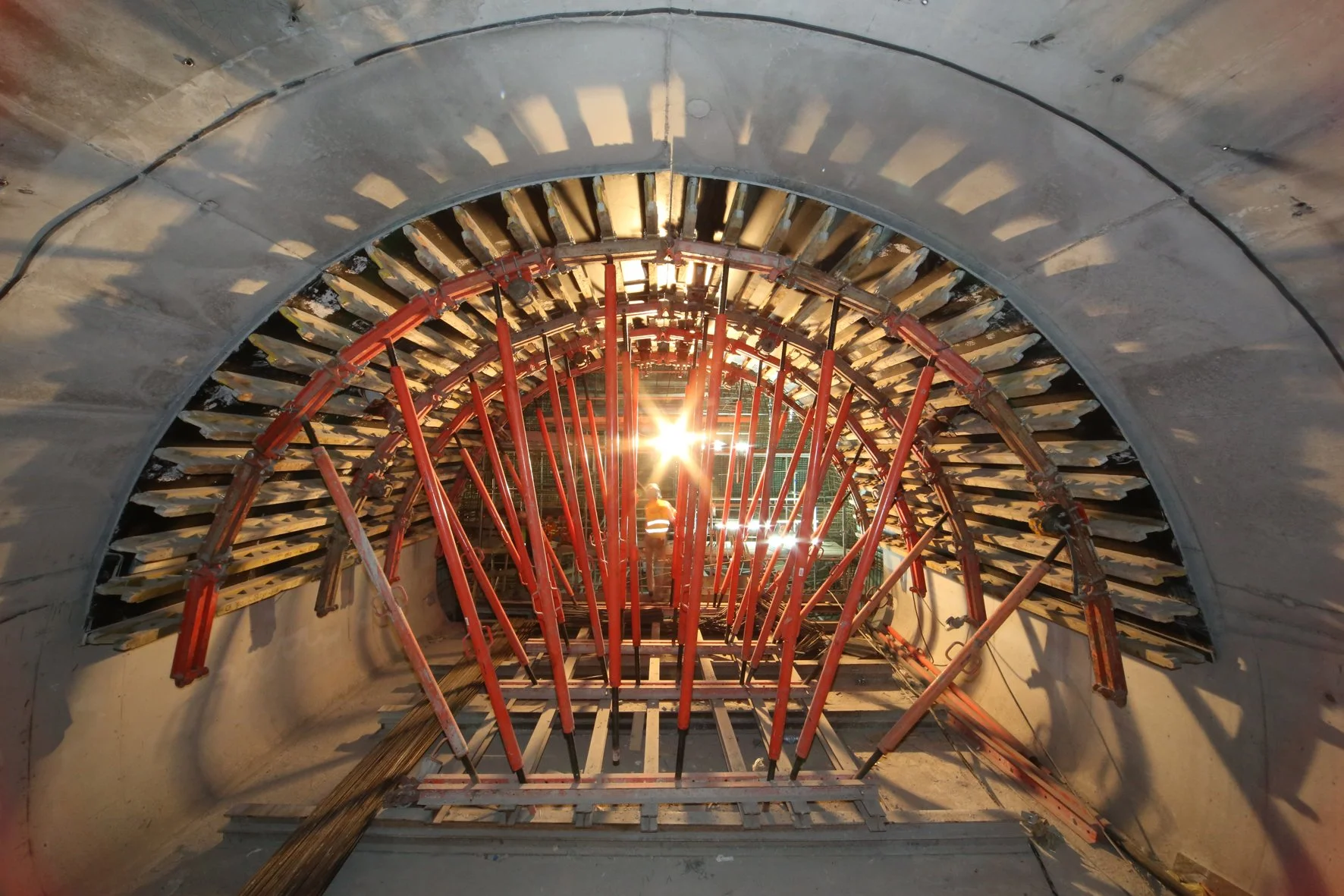 View inside a tunnel under construction with red scaffolding and concrete walls, sunlight shining through the opening.