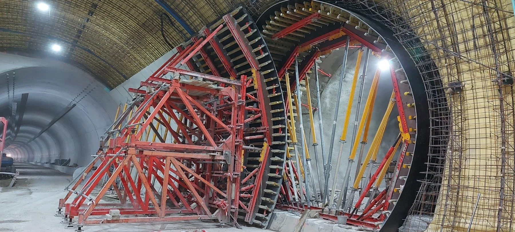 Construction of a large tunnel with various scaffolding and support structures, including red, yellow, and metal beams, inside an arched tunnel bore.