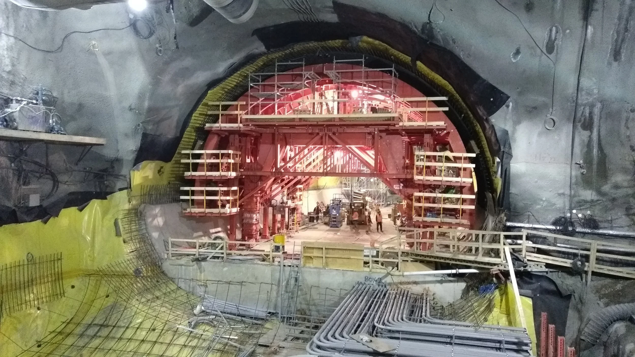 Underground tunnel construction site with scaffolding, workers, and construction equipment. The tunnel is being excavated with visible rebar, pipes, and safety barriers.