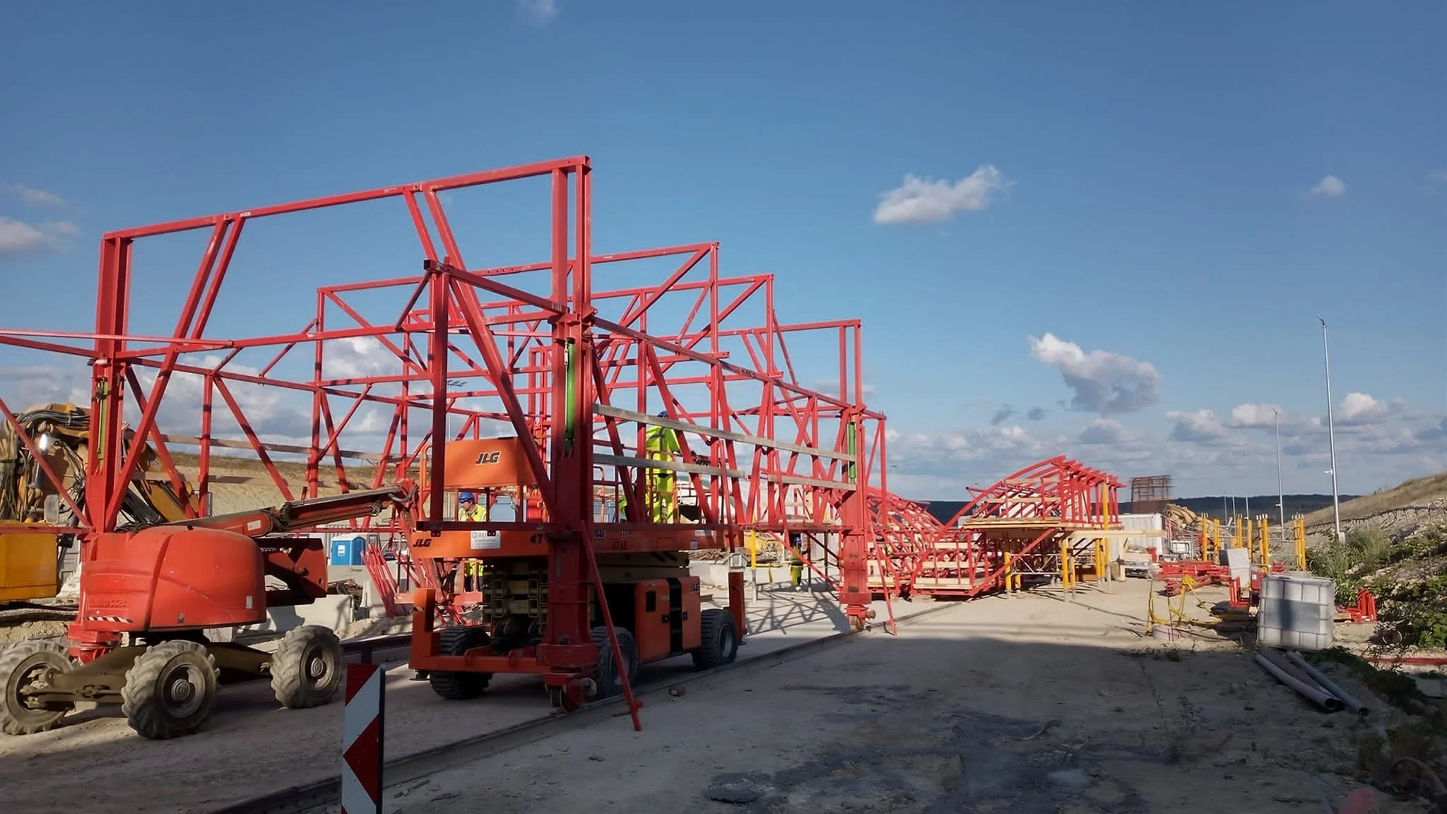 Construction site with red metal framework, scaffolding, and equipment under a blue sky with scattered clouds.