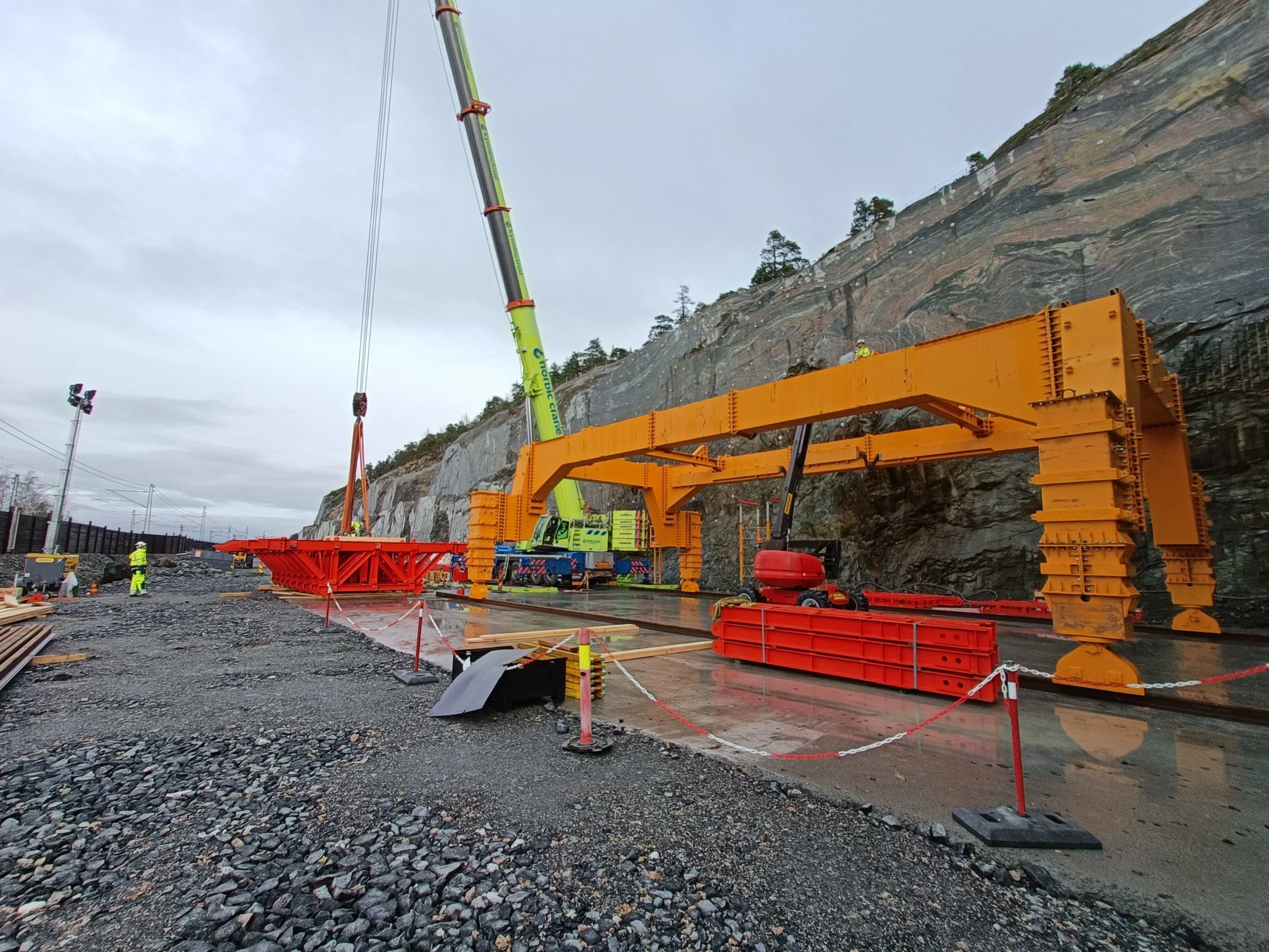 Construction site with large orange steel framework, crane, and workers on a rocky outdoor area next to a cliff face.
