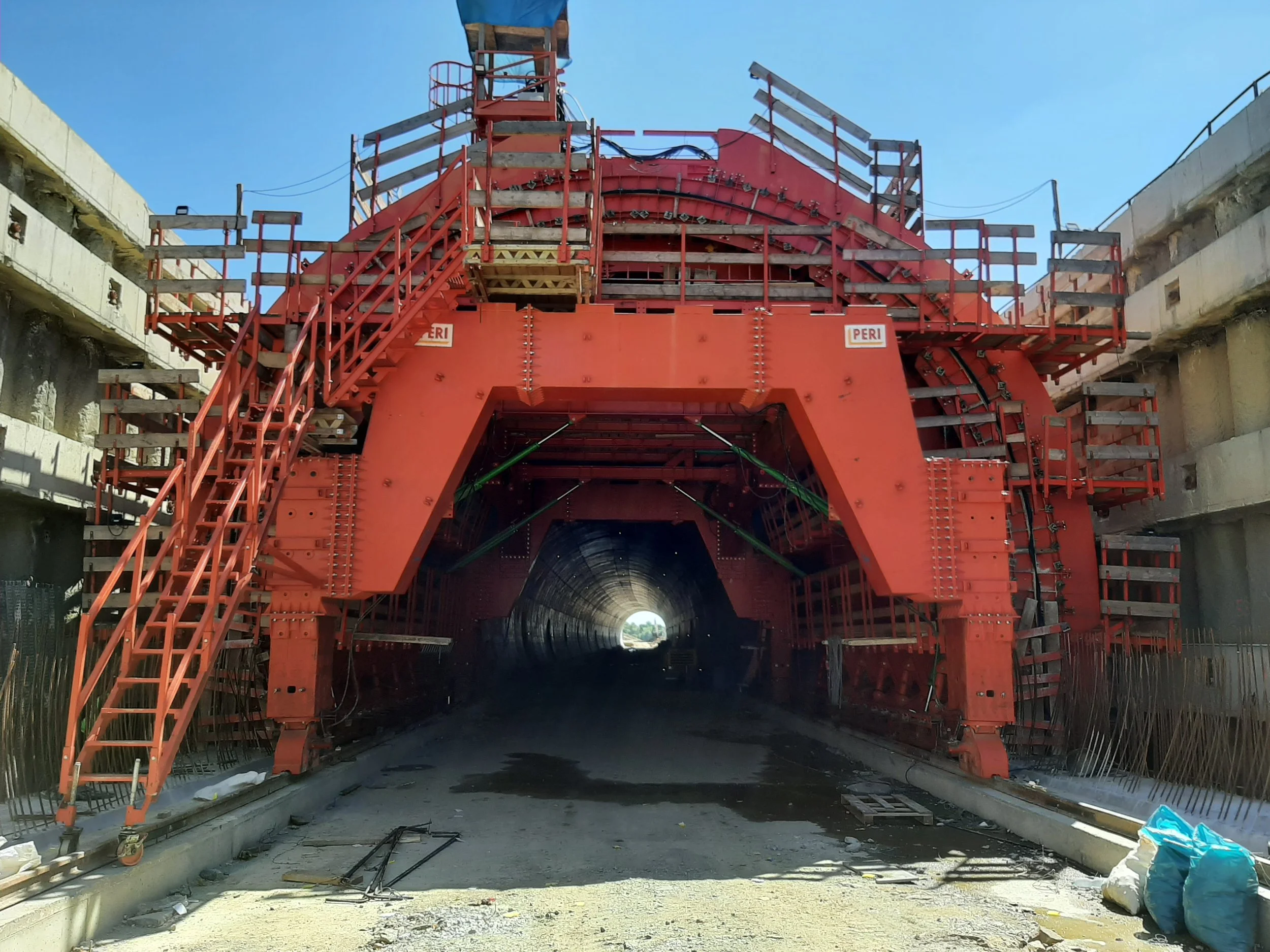 Construction site with a large red tunnel boring machine and surrounding concrete structures under a blue sky.