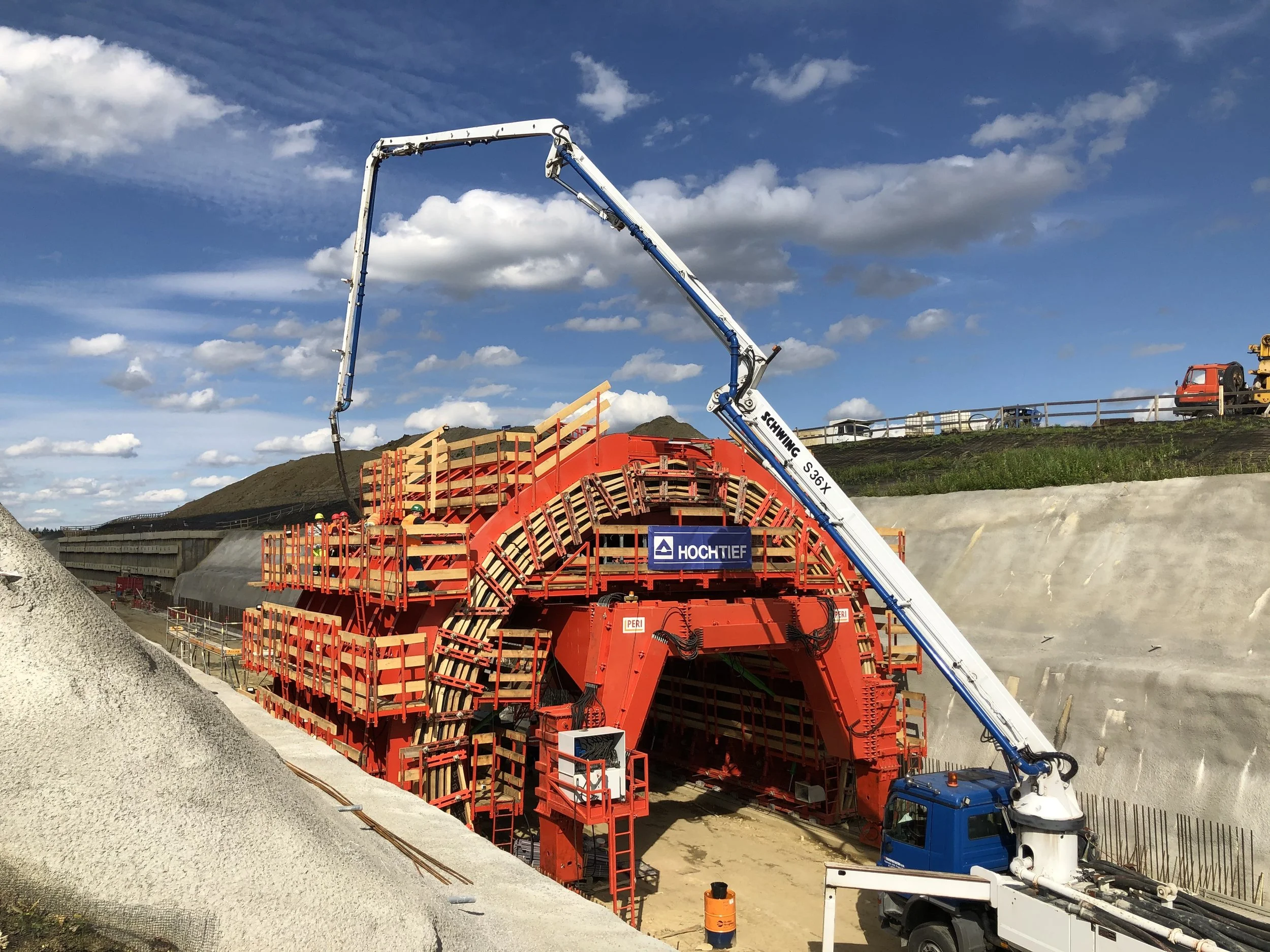 Construction site with a tunnel boring machine in operation, orange and red machinery tunnel entrance, blue sky with clouds in the background.