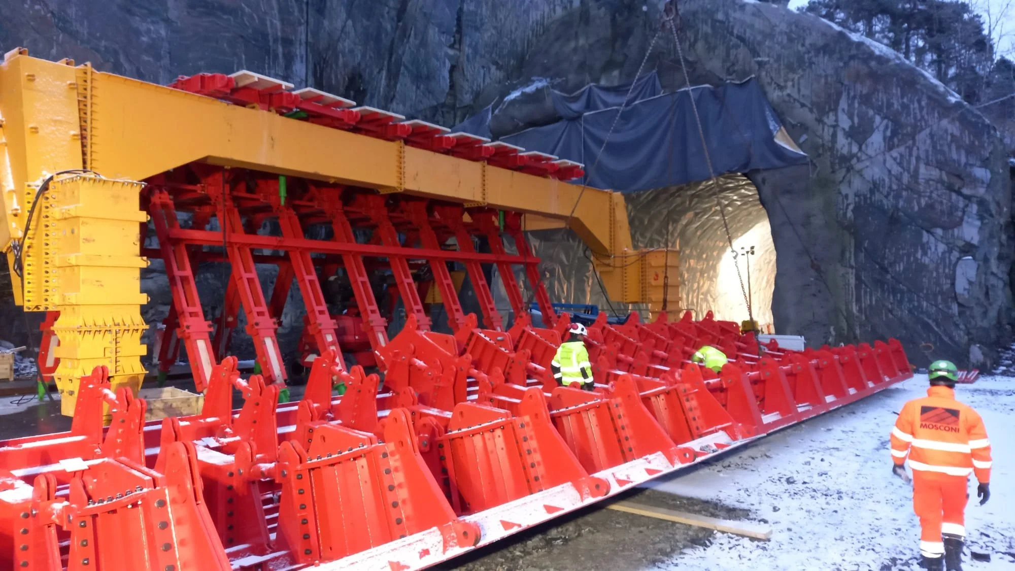 Workers in high-visibility clothing inspecting a large tunnel excavation using heavy machinery with orange ground protection barriers and exposed rock walls.