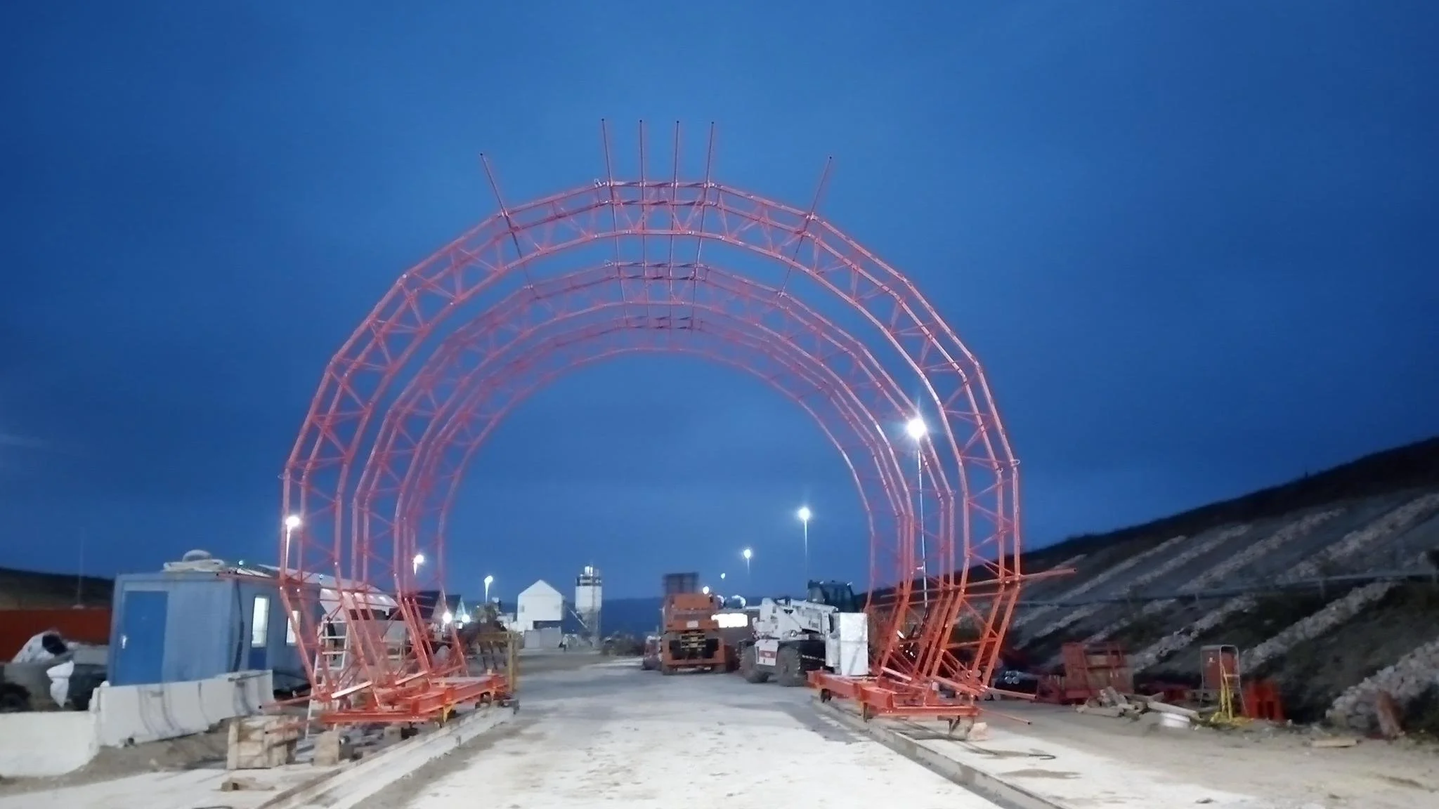 Under construction site at dusk with a large red metal arch framework and construction vehicles.