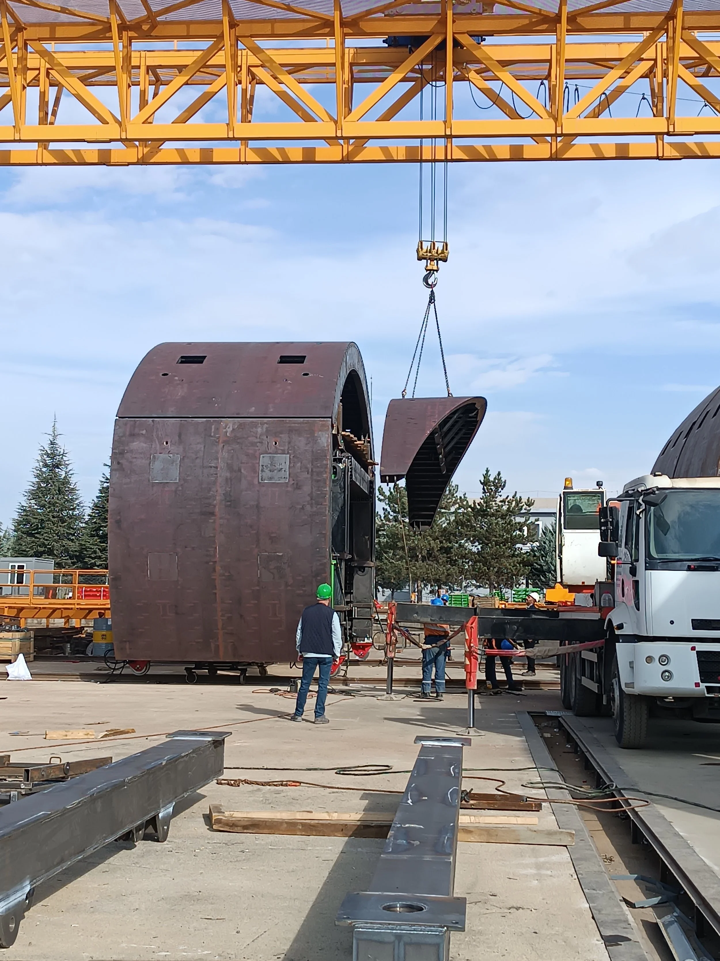 Construction site with a yellow crane lifting a large curved steel piece to install into a large, curved structure, with workers guiding the process.