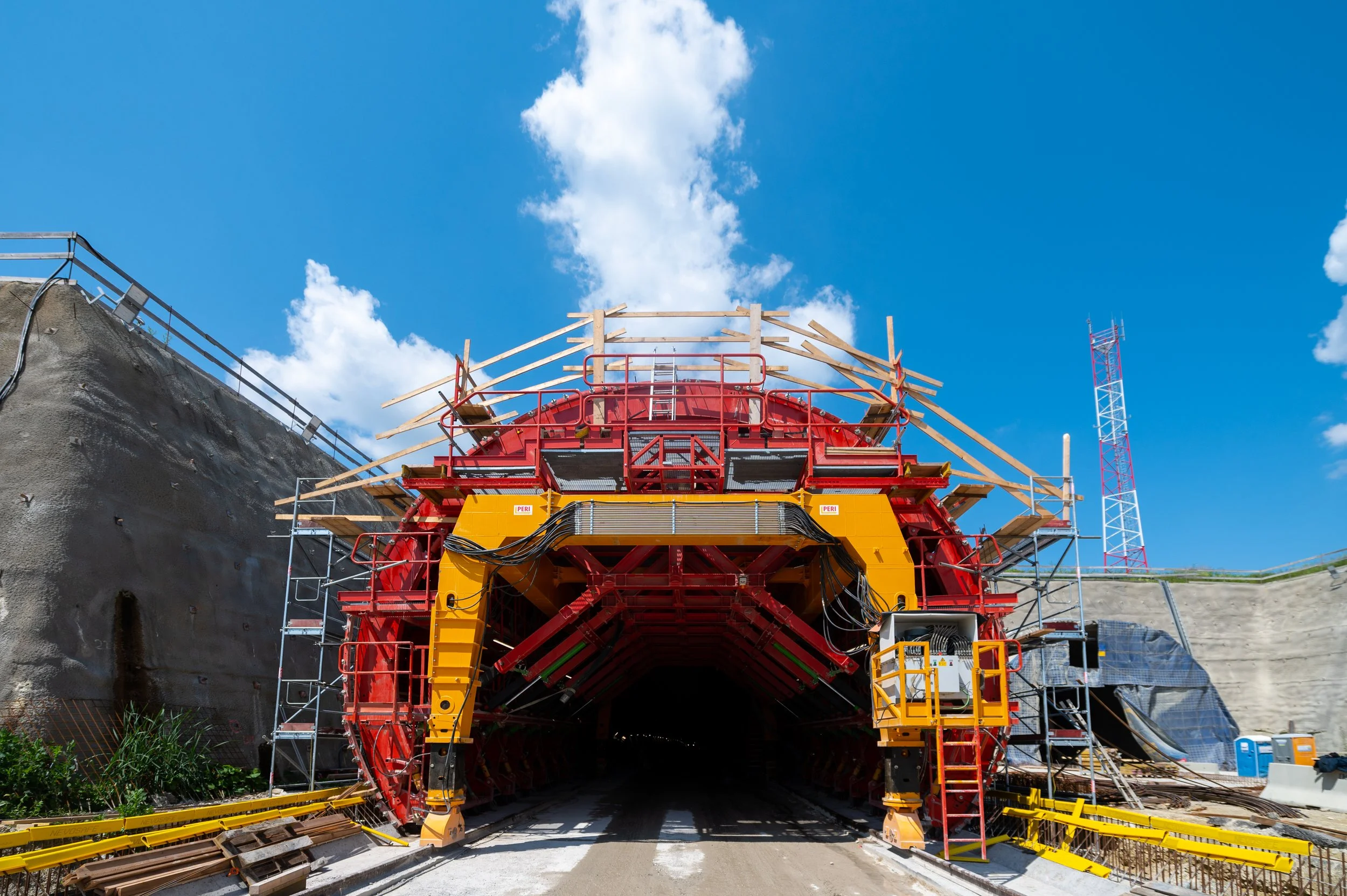 Construction tunnel under construction with scaffolding and support structures, blue sky with clouds in the background.