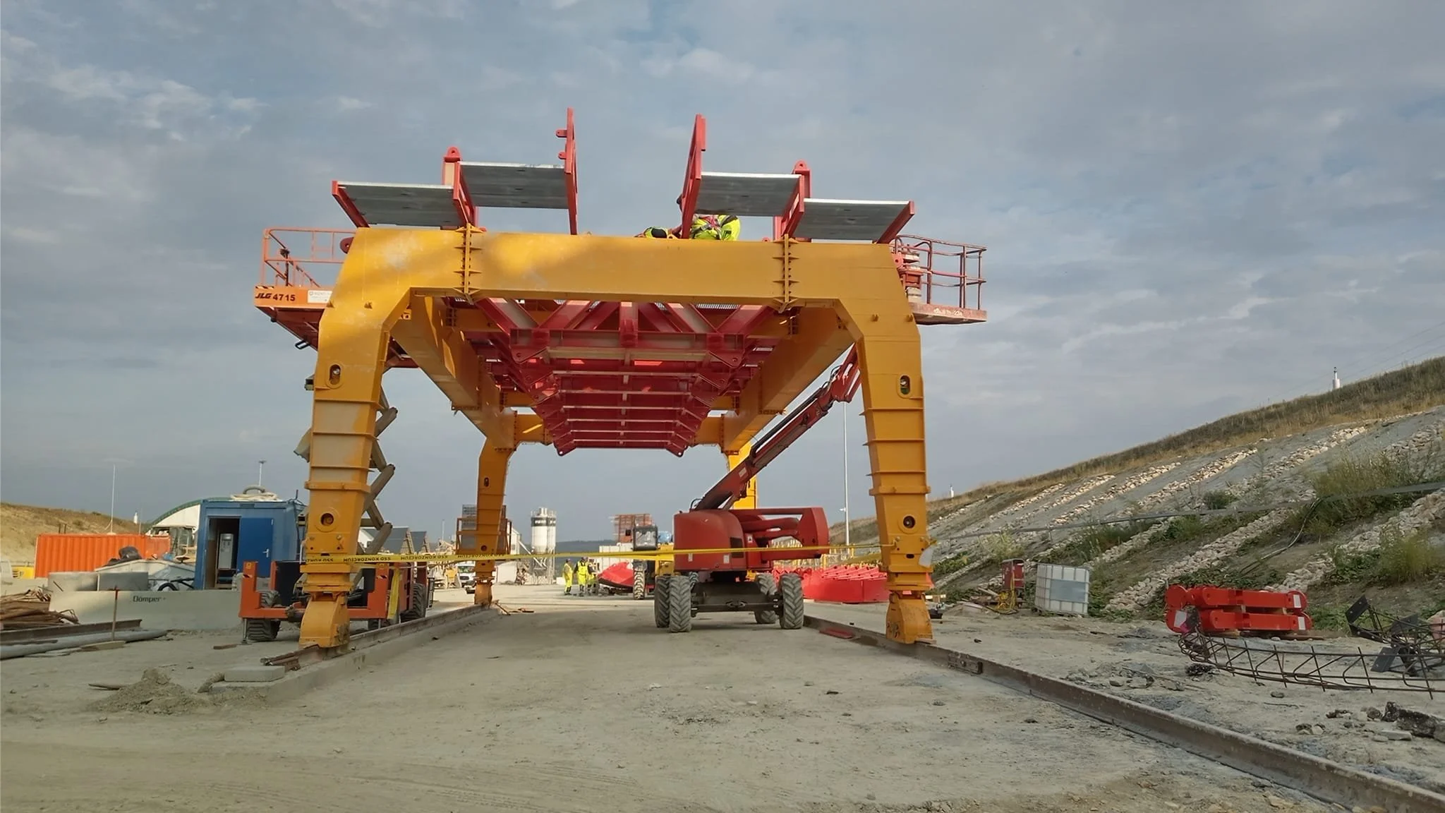 Construction site with large yellow and red machinery, workers in yellow safety vests, and a cloudy sky.